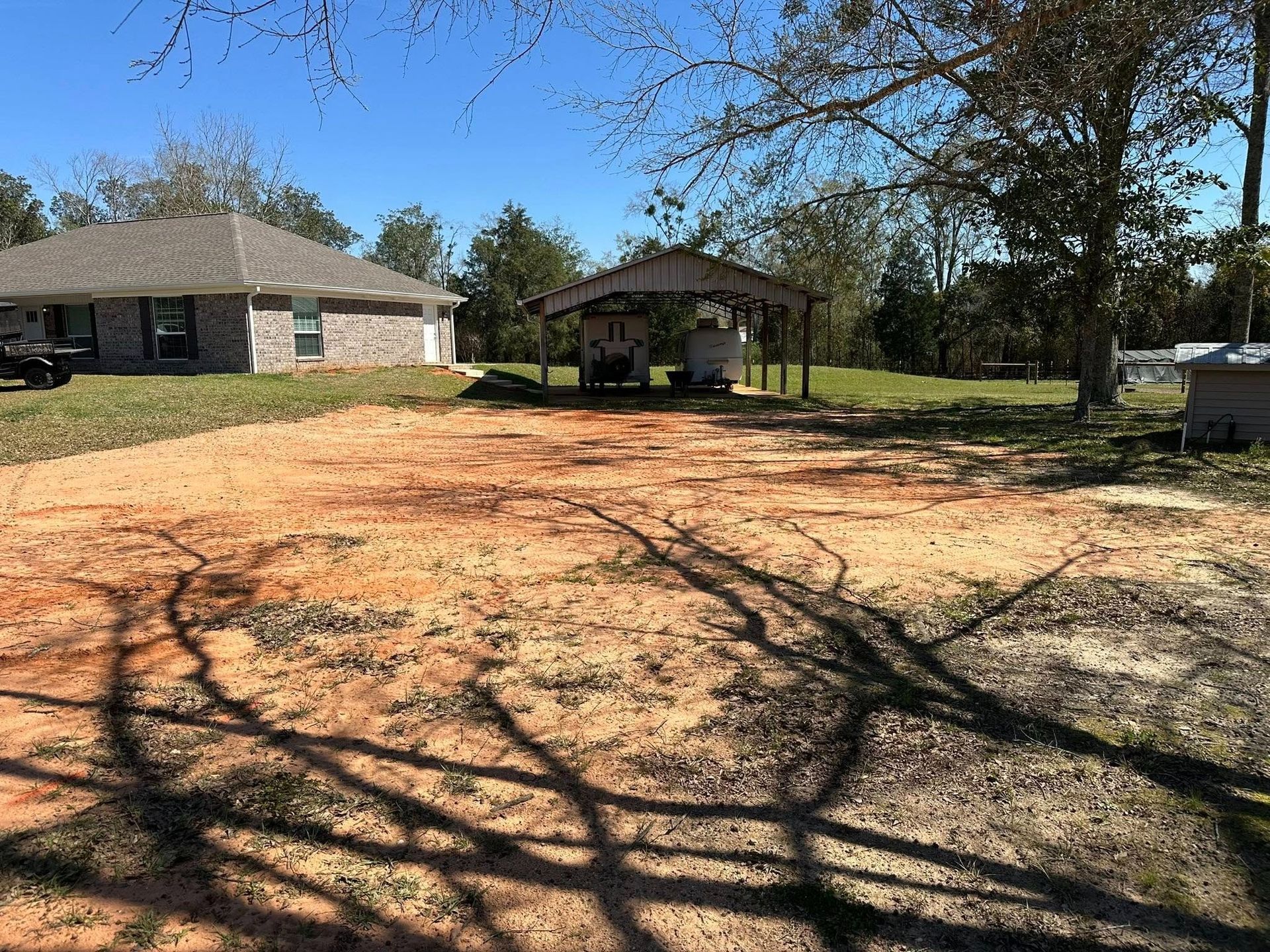 Dirt yard with carport, house in the background under a blue sky. Sunlight casts shadows.