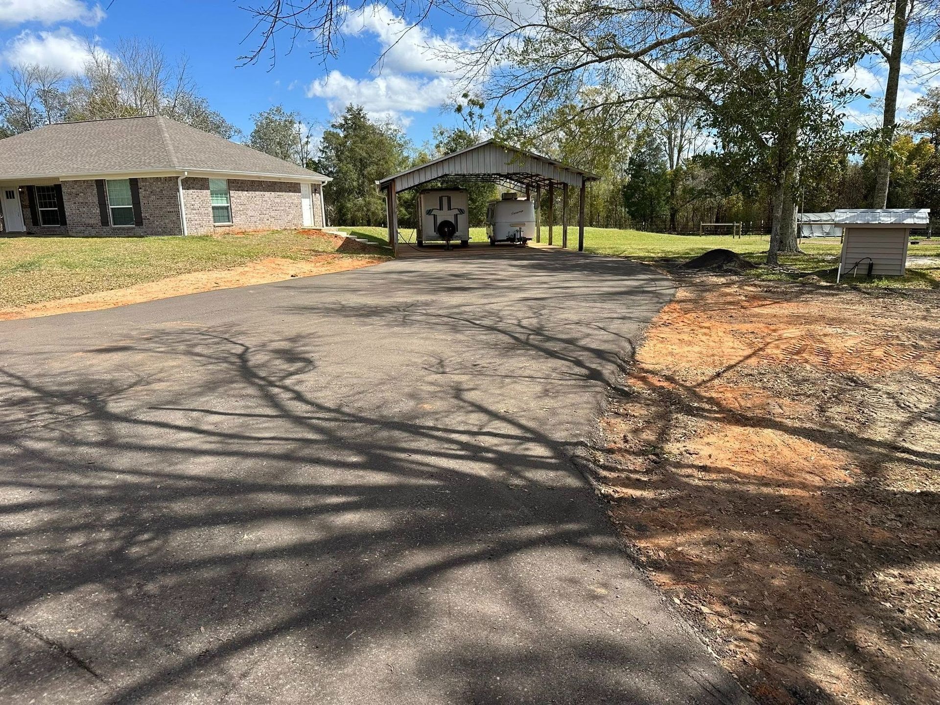 A paved driveway leads to a carport and house; trees frame the background, and blue sky with clouds.