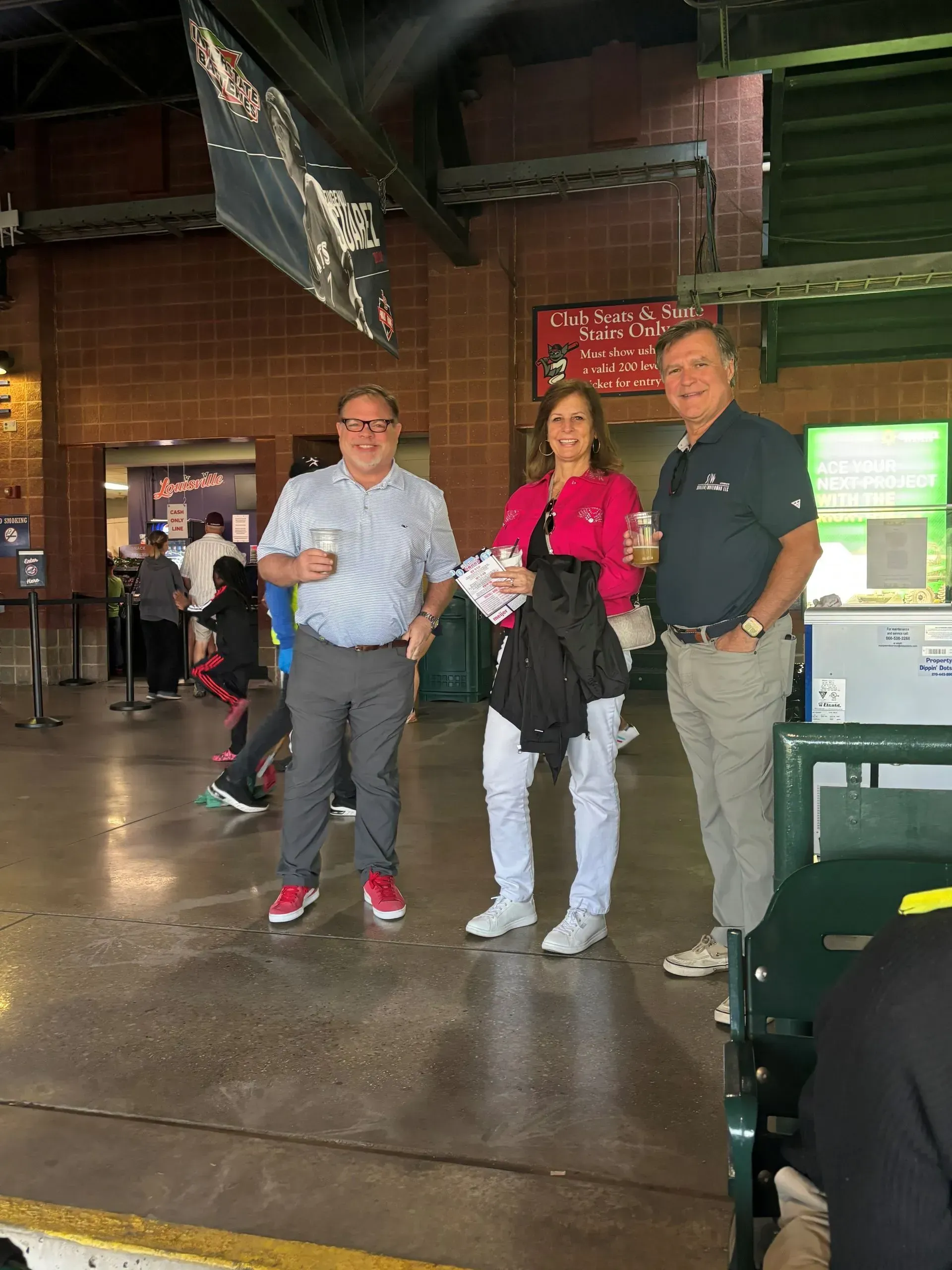 Three people at a baseball stadium: two men and a woman. They are holding drinks and smiling.