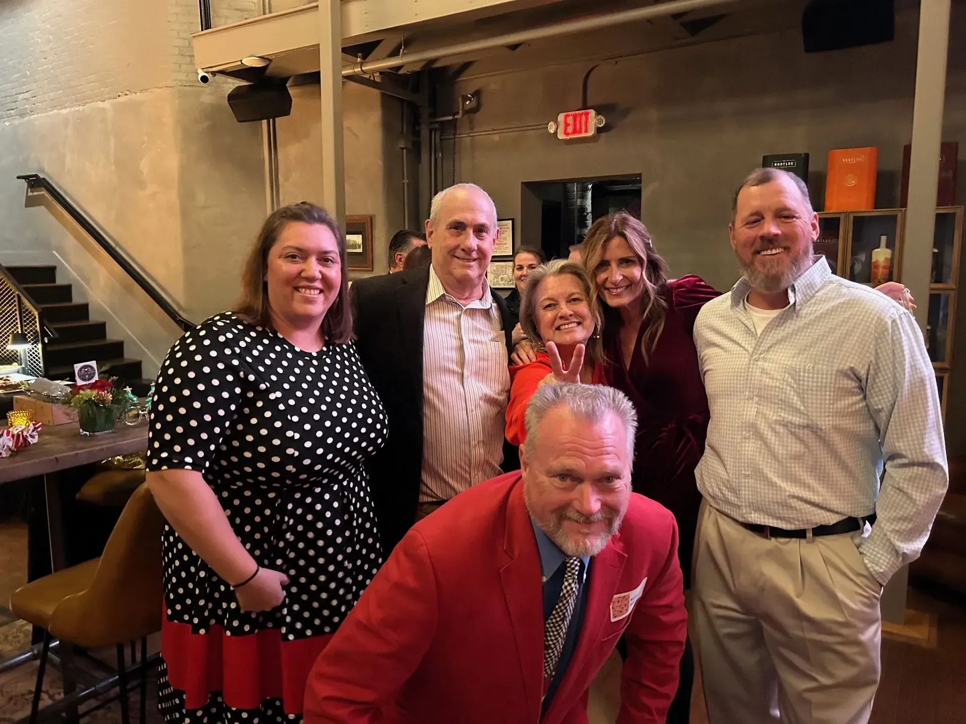 Group of six people smiling at a gathering inside a building. One man wears a red jacket.