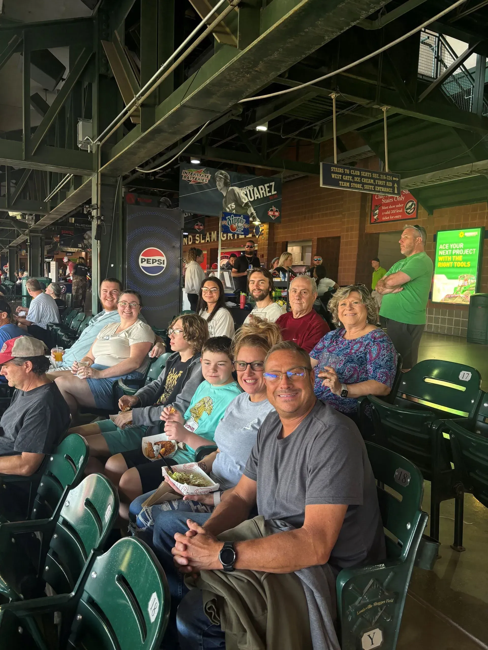 Group of people at a baseball game, smiling, seated in green stadium chairs, under an overhang.