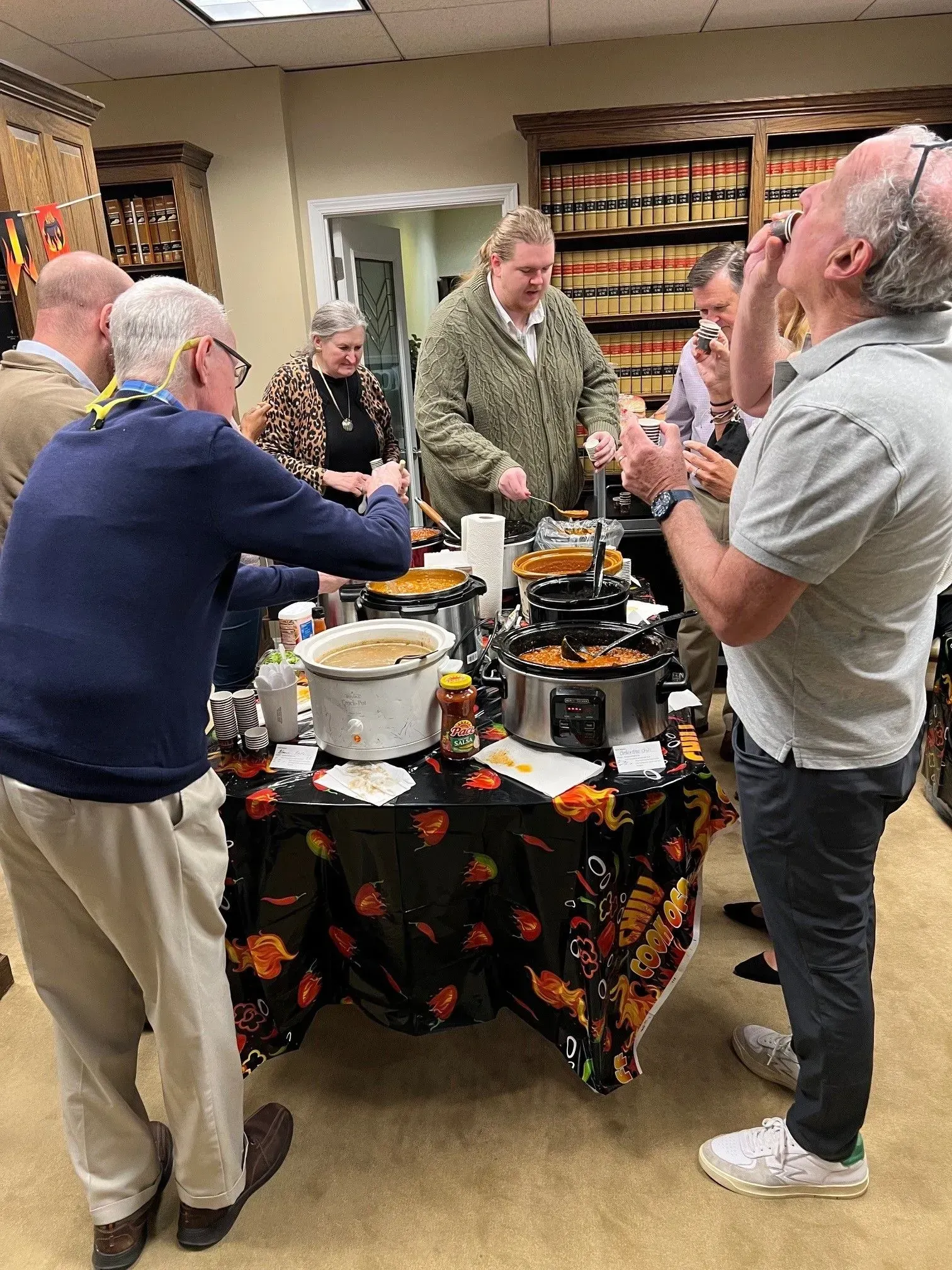 People gathered around a table with slow cookers. Someone serves food. Indoors, fall-themed tablecloth.
