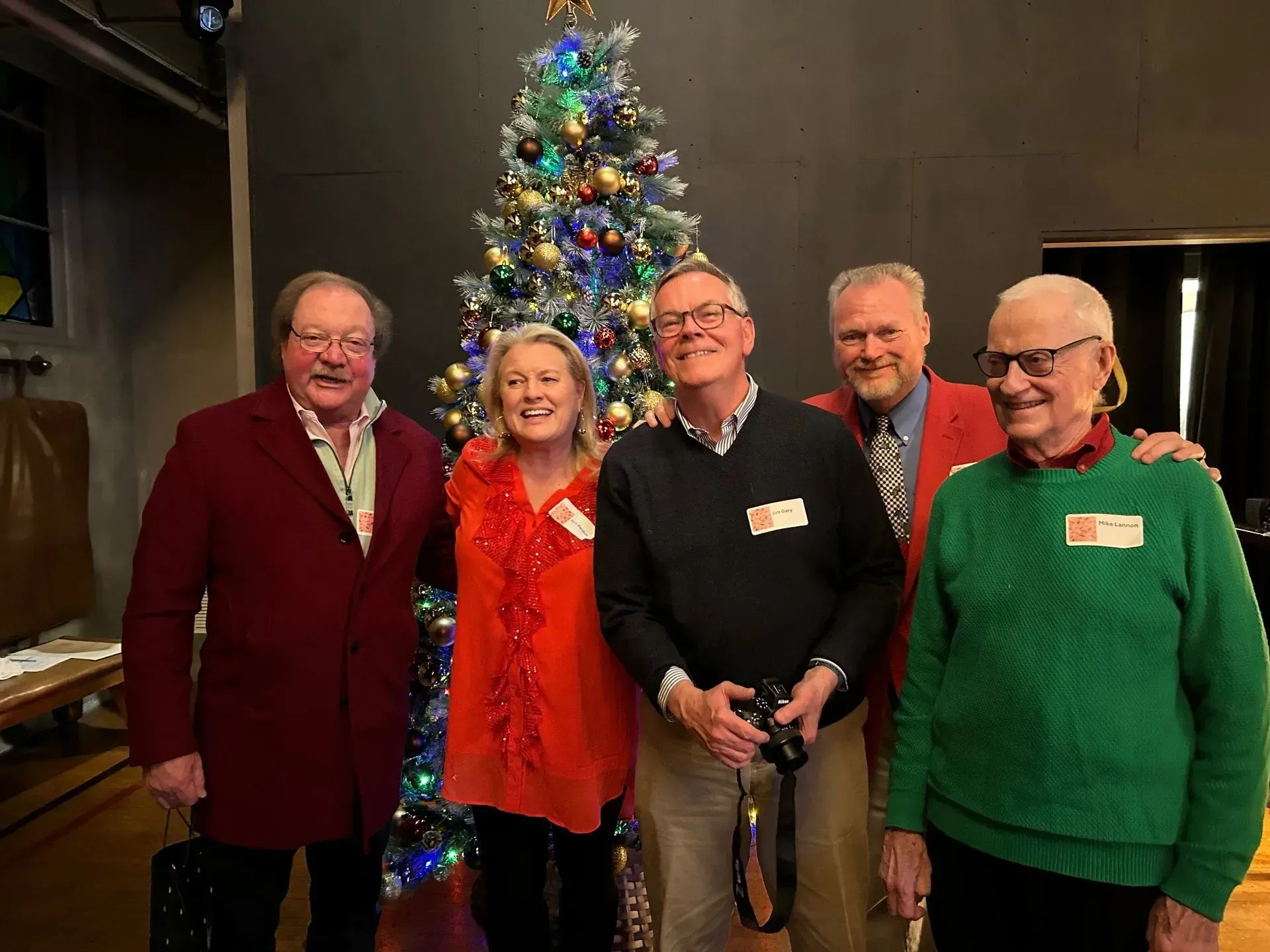 Five people smiling by a Christmas tree. Two men in red coats, one in green sweater. Woman in orange. Everyone wearing nametags.