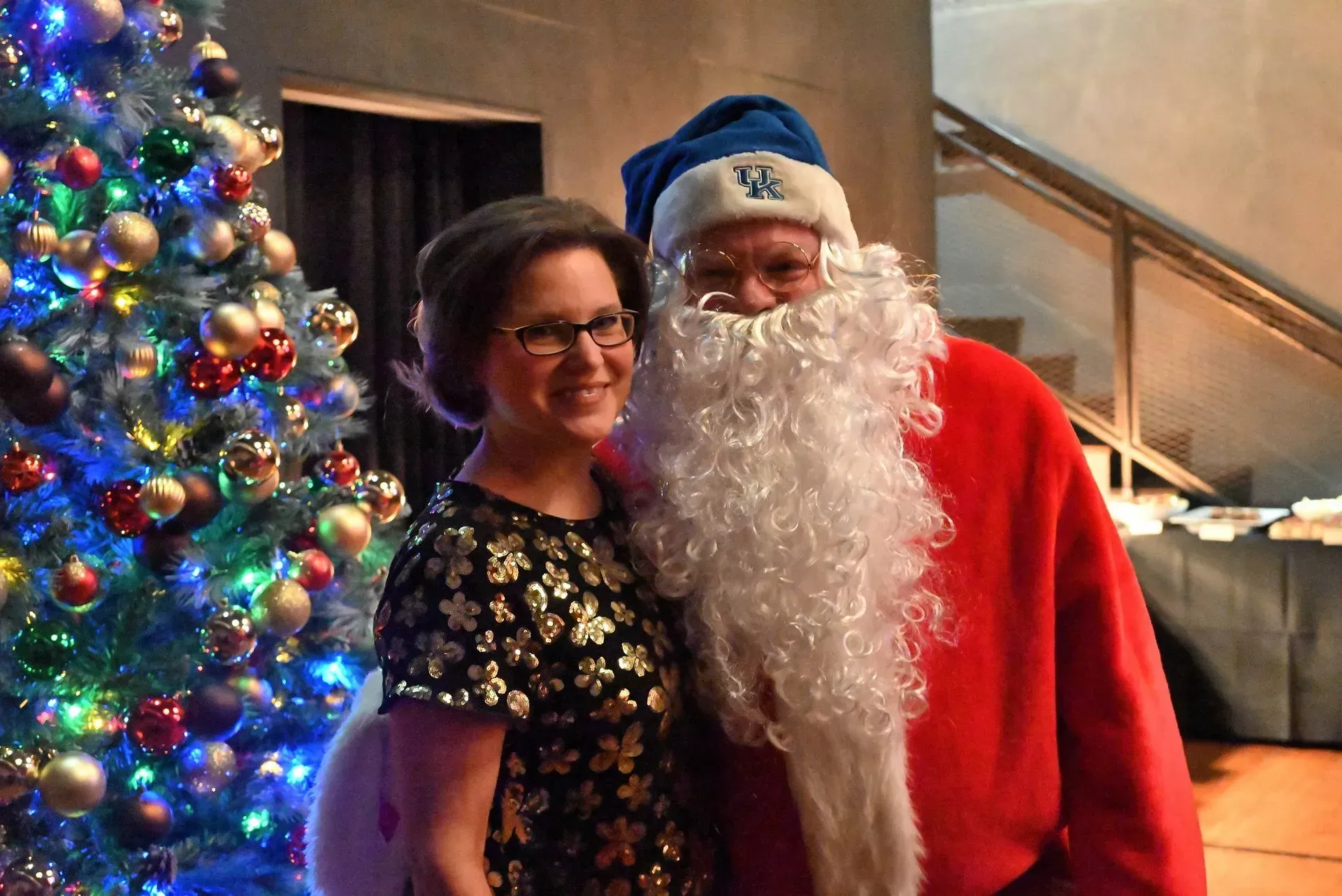 Woman poses with Santa Claus in front of a Christmas tree, both smiling. Santa wears a blue UK hat.