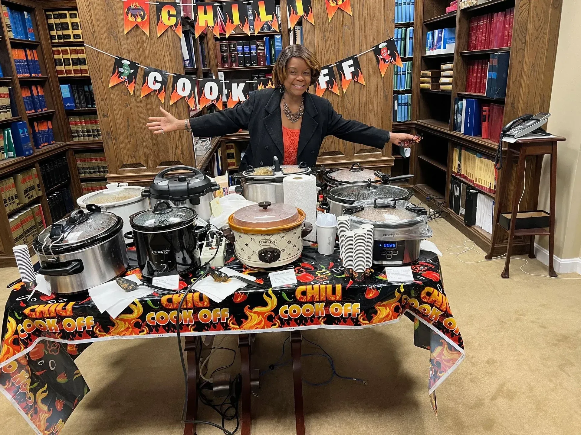 Woman with arms open at a chili cook-off table with many slow cookers and festive decor.