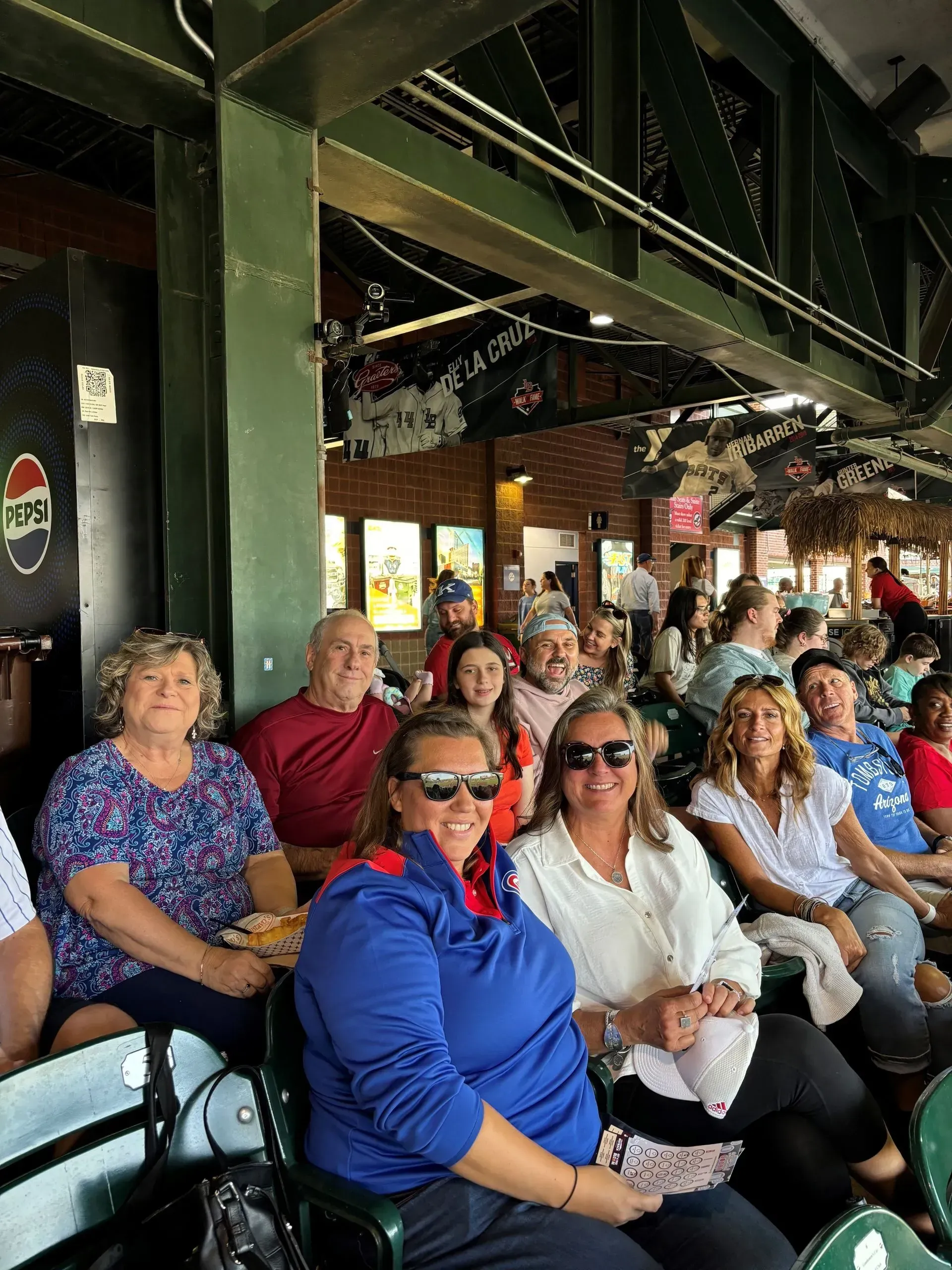 Group of people at a baseball game, under a green support beam. Some smiling, wearing casual clothes.