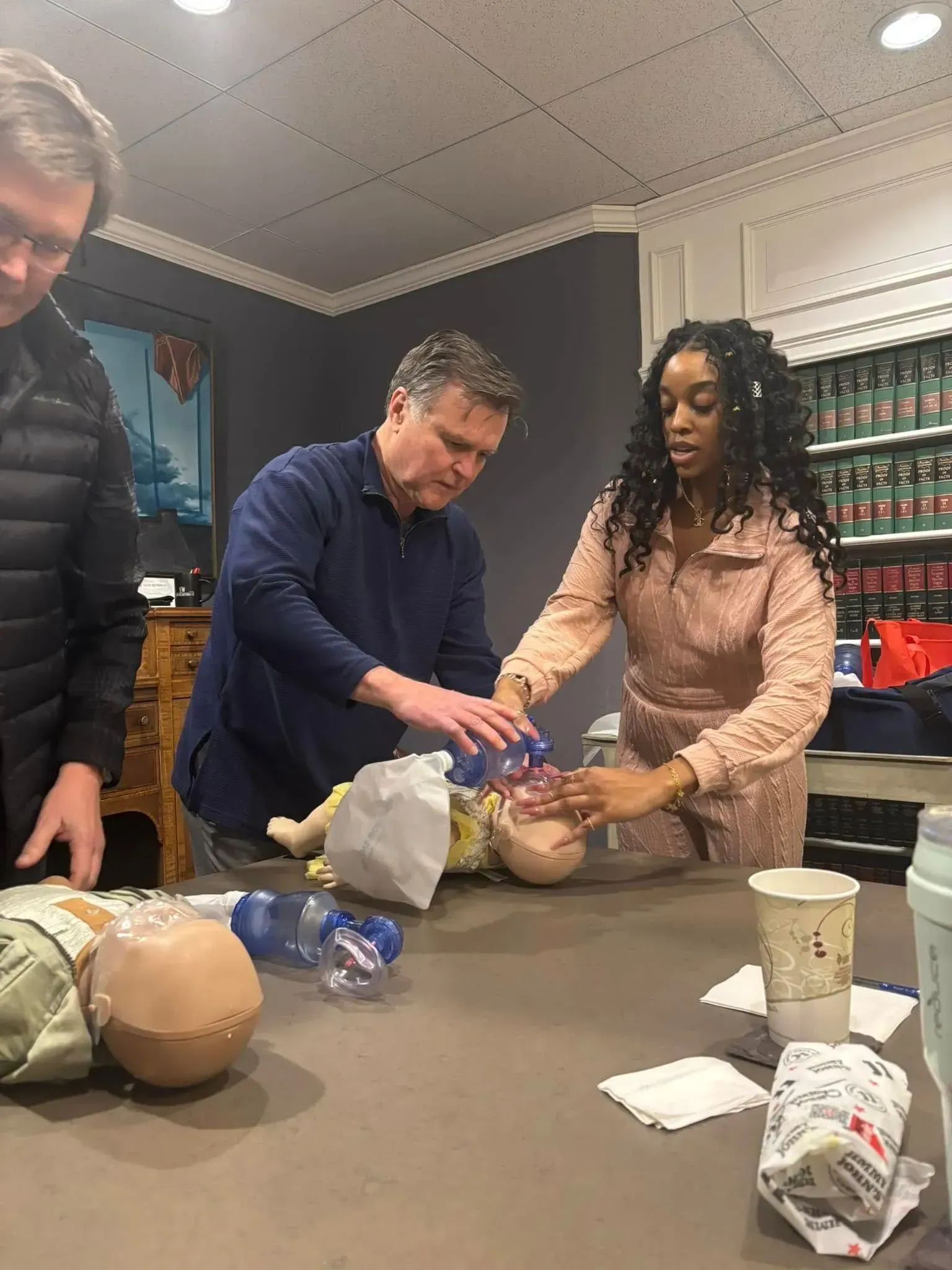 Three people practice baby CPR on a training mannequin. They are at a table indoors.