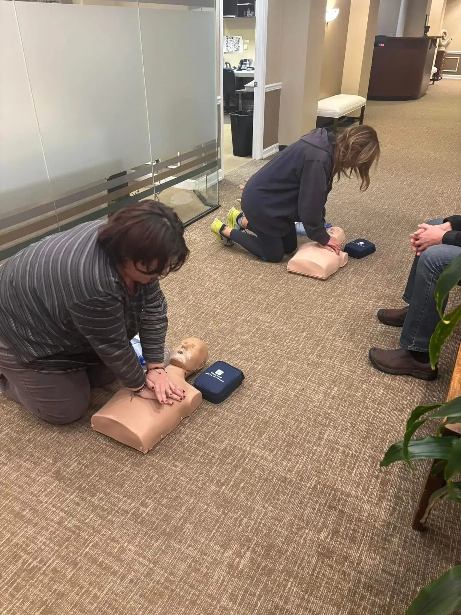 Two people practice CPR on mannequins during a training session, office setting.