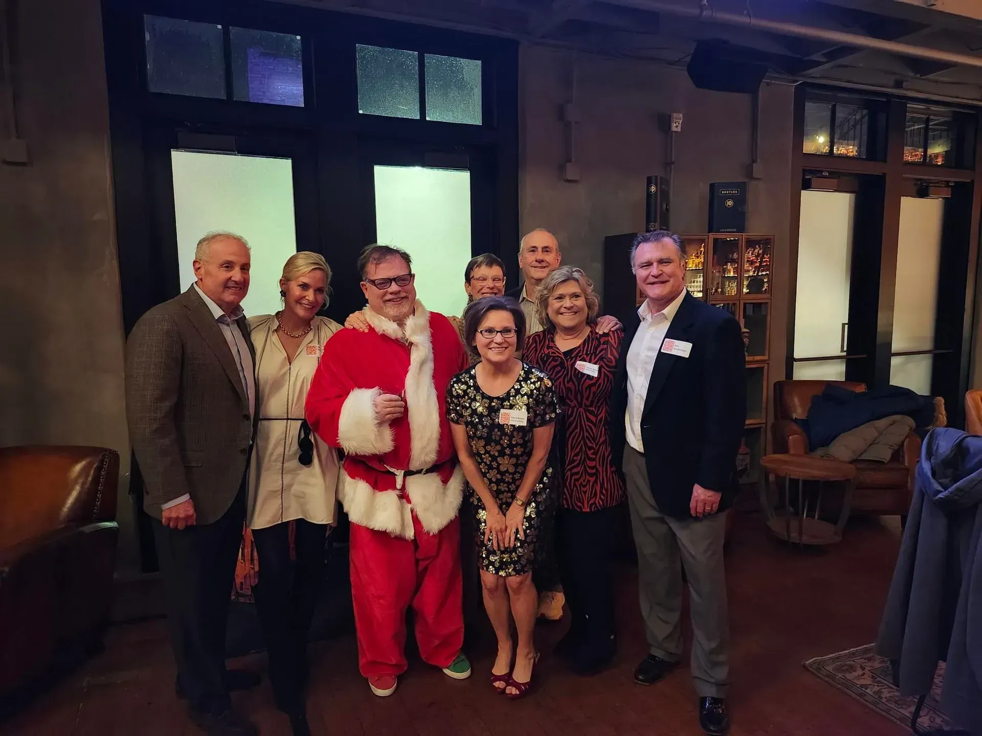 Group of people at a holiday party, including someone dressed as Santa, in a warmly lit interior.