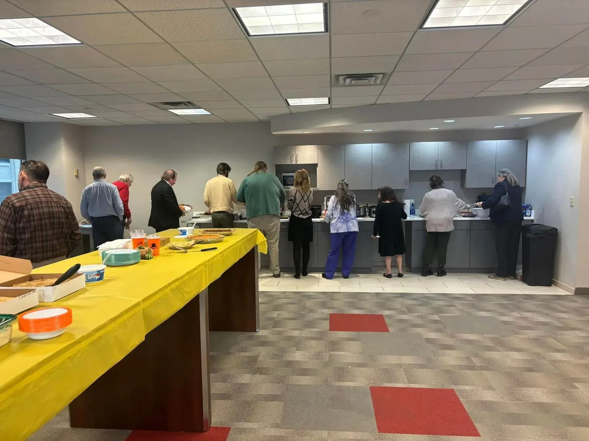 People in an office kitchen serving themselves from a buffet, long yellow table in front.