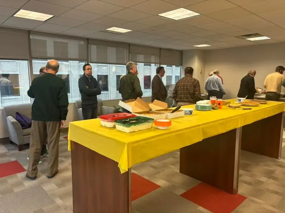 People at a casual office gathering with food on a yellow table. Windows and a couch are visible.