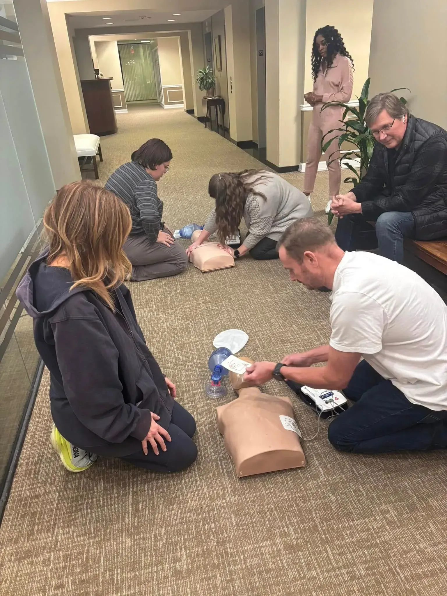 People practicing CPR on mannequins in a hallway, with an instructor.