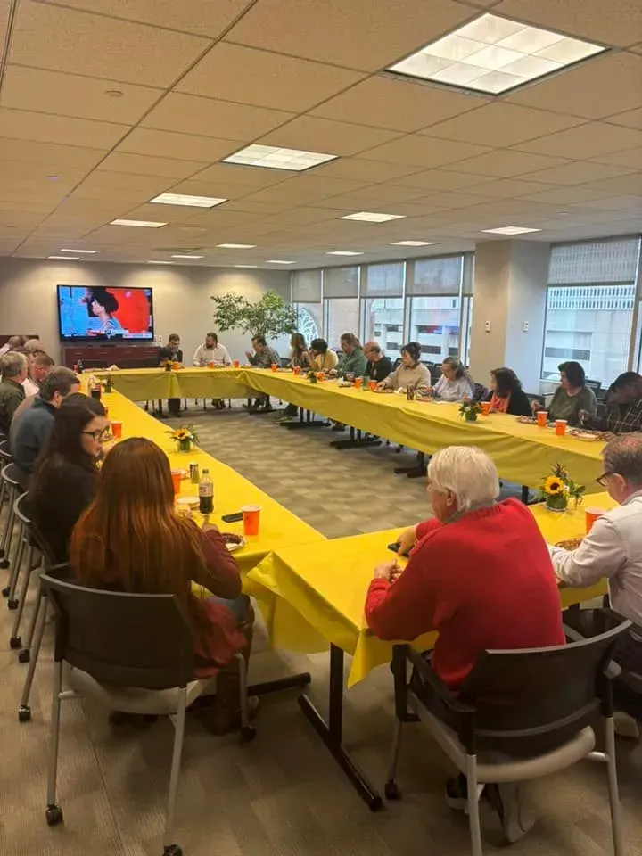 People seated at a long, yellow-covered table in a well-lit office, watching a screen.