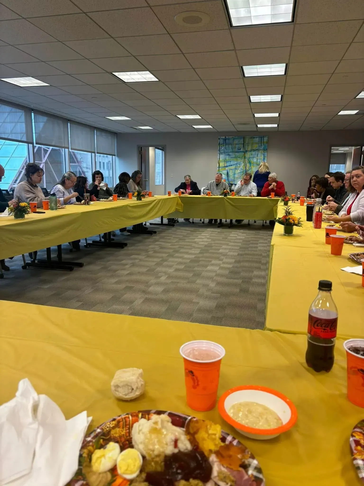 People seated at a long table with yellow tablecloths, enjoying a meal. Indoors with large windows.