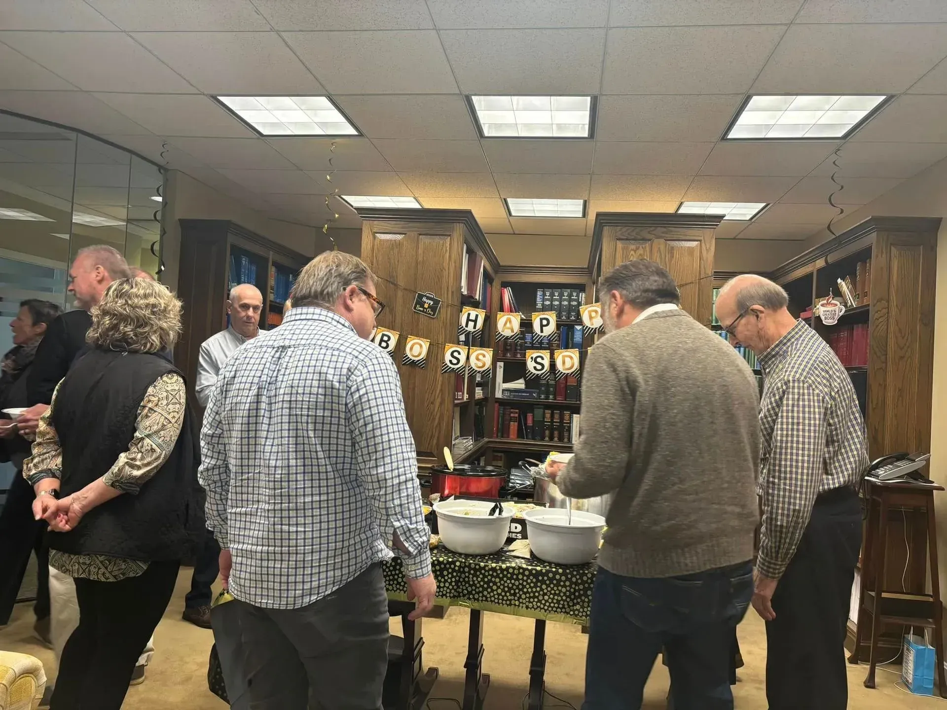 Group of people at a gathering, some gathered around a table with food in an office setting.