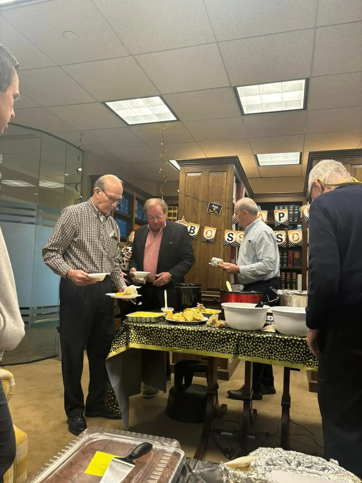 Men at a party: eating food from a buffet table, indoor setting, one man in apron, others holding plates.