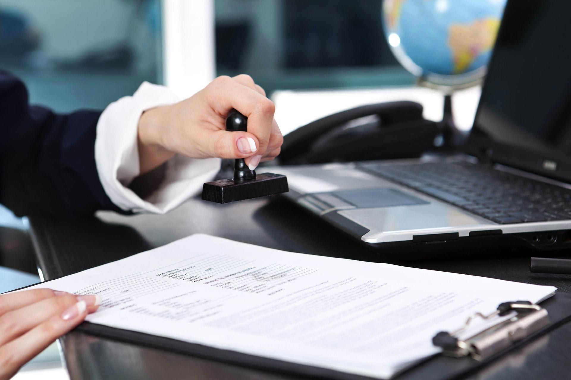A person in a business suit stamps a document on a desk with a laptop and globe in the background.