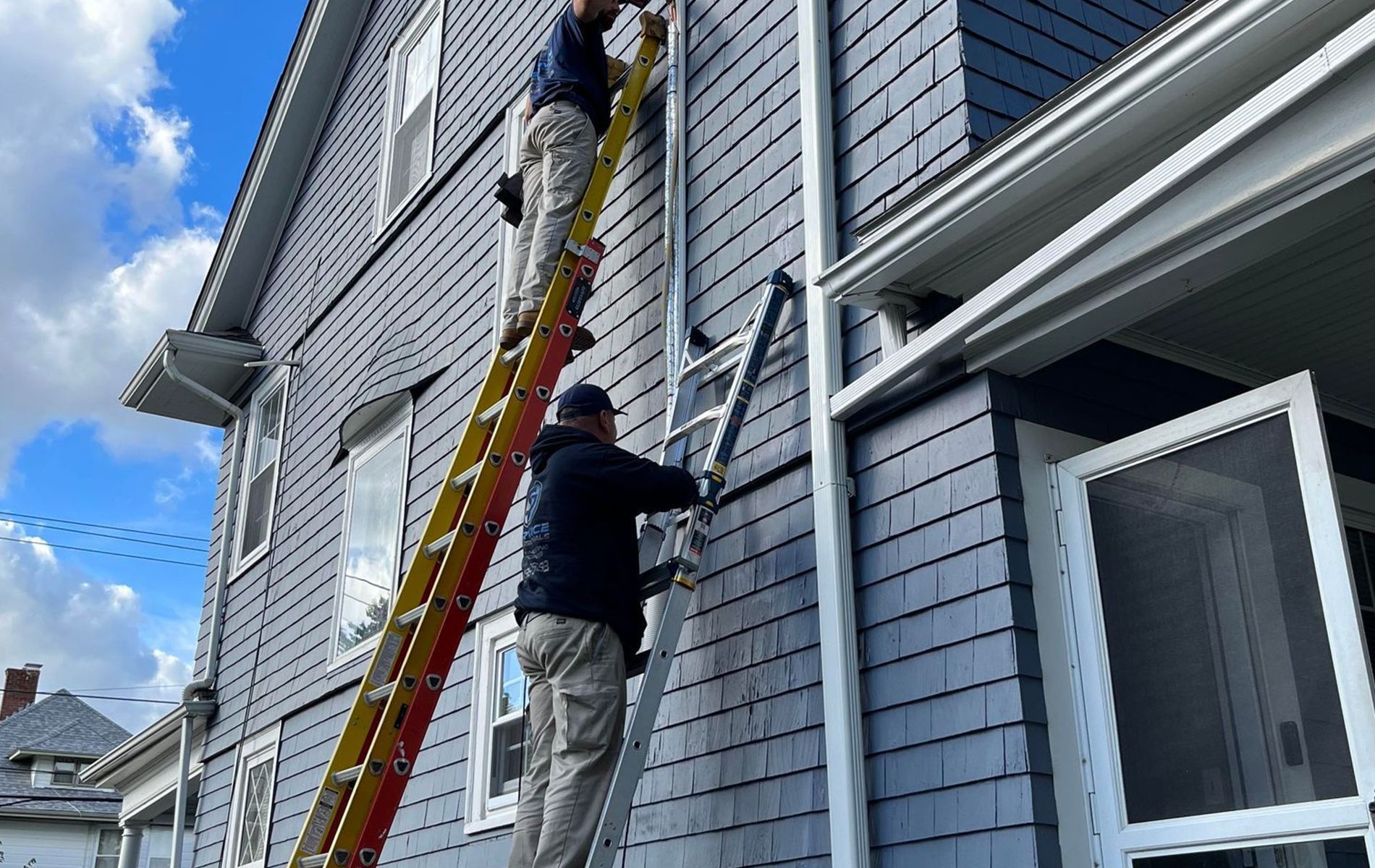 Two people on ladders working on the side of a gray house with white trim.