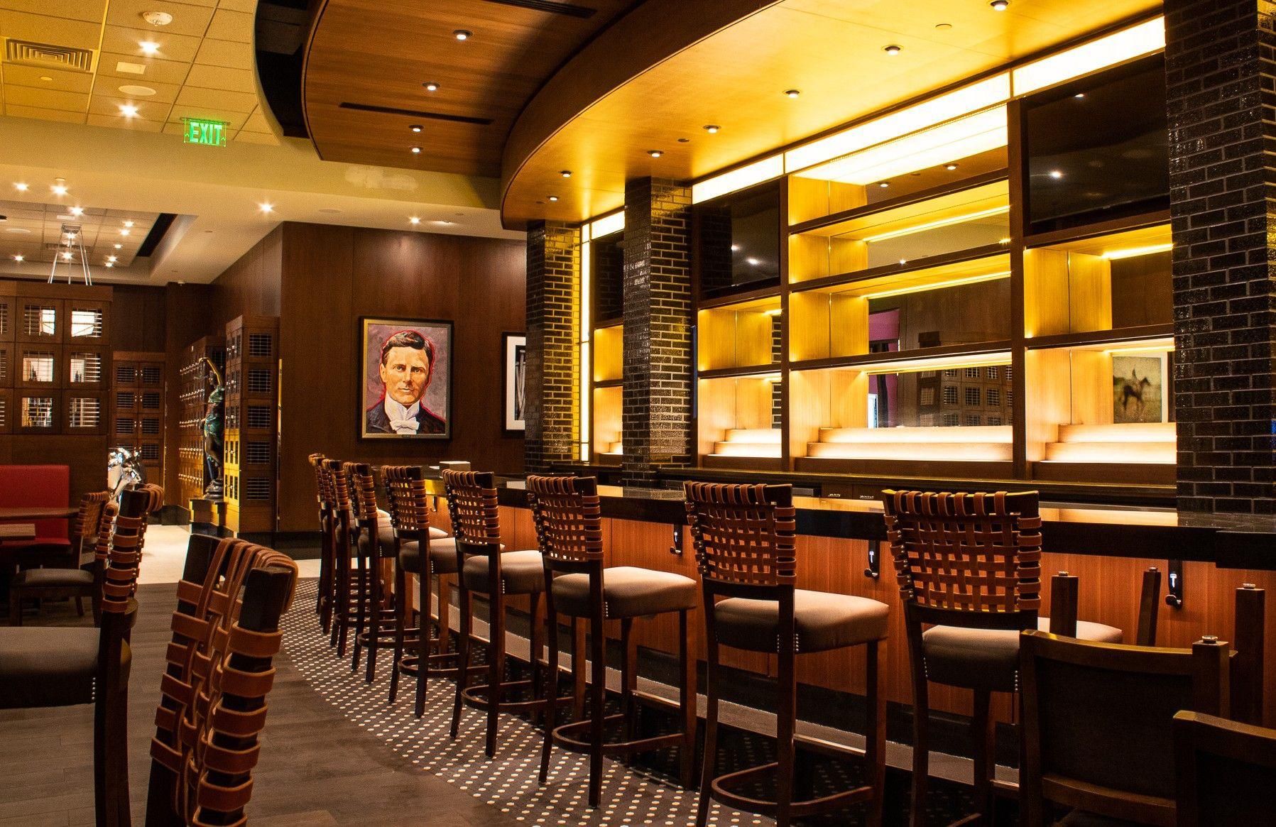Warm, upscale bar interior with rows of stools, glowing shelves, and framed wall art.