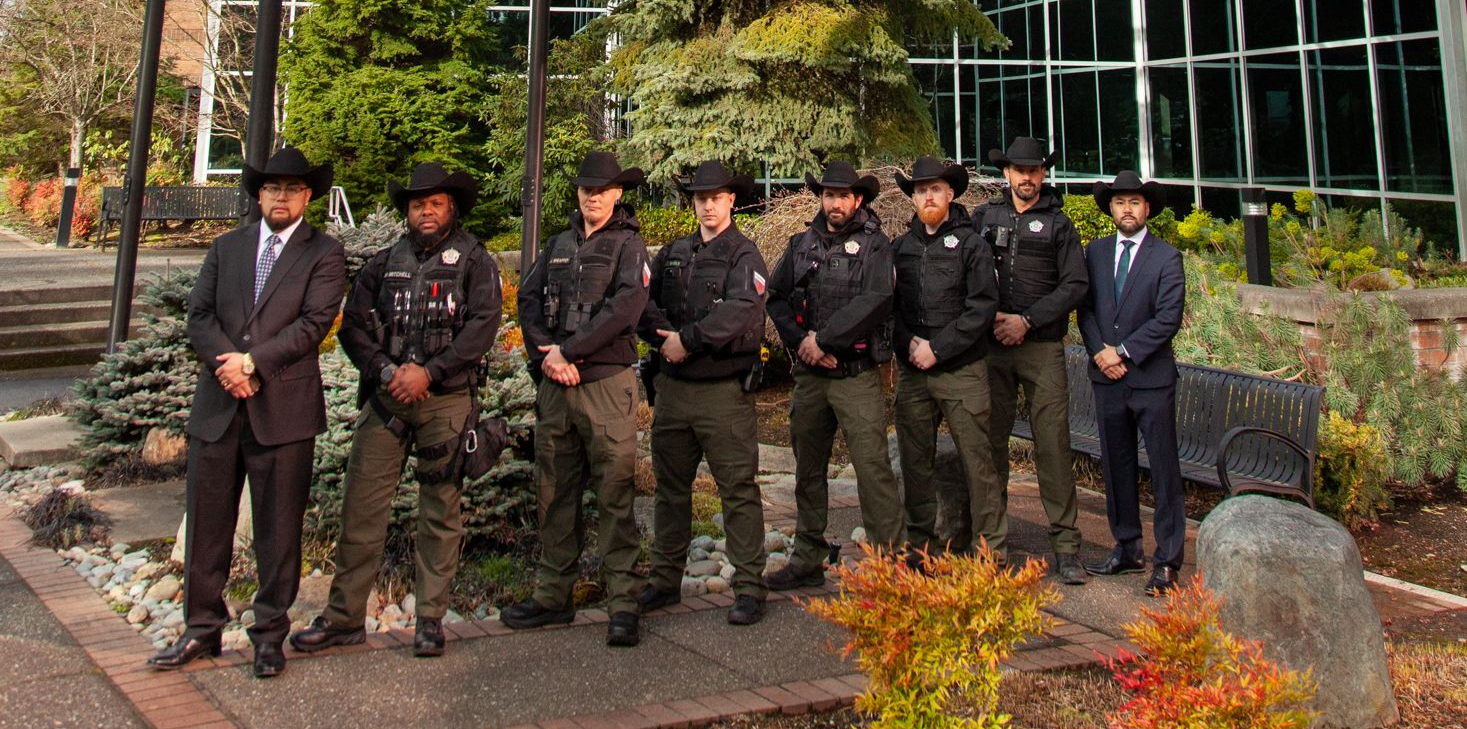 Eight uniformed officers stand in a line before a modern glass building and three flagpoles under a cloudy sky.