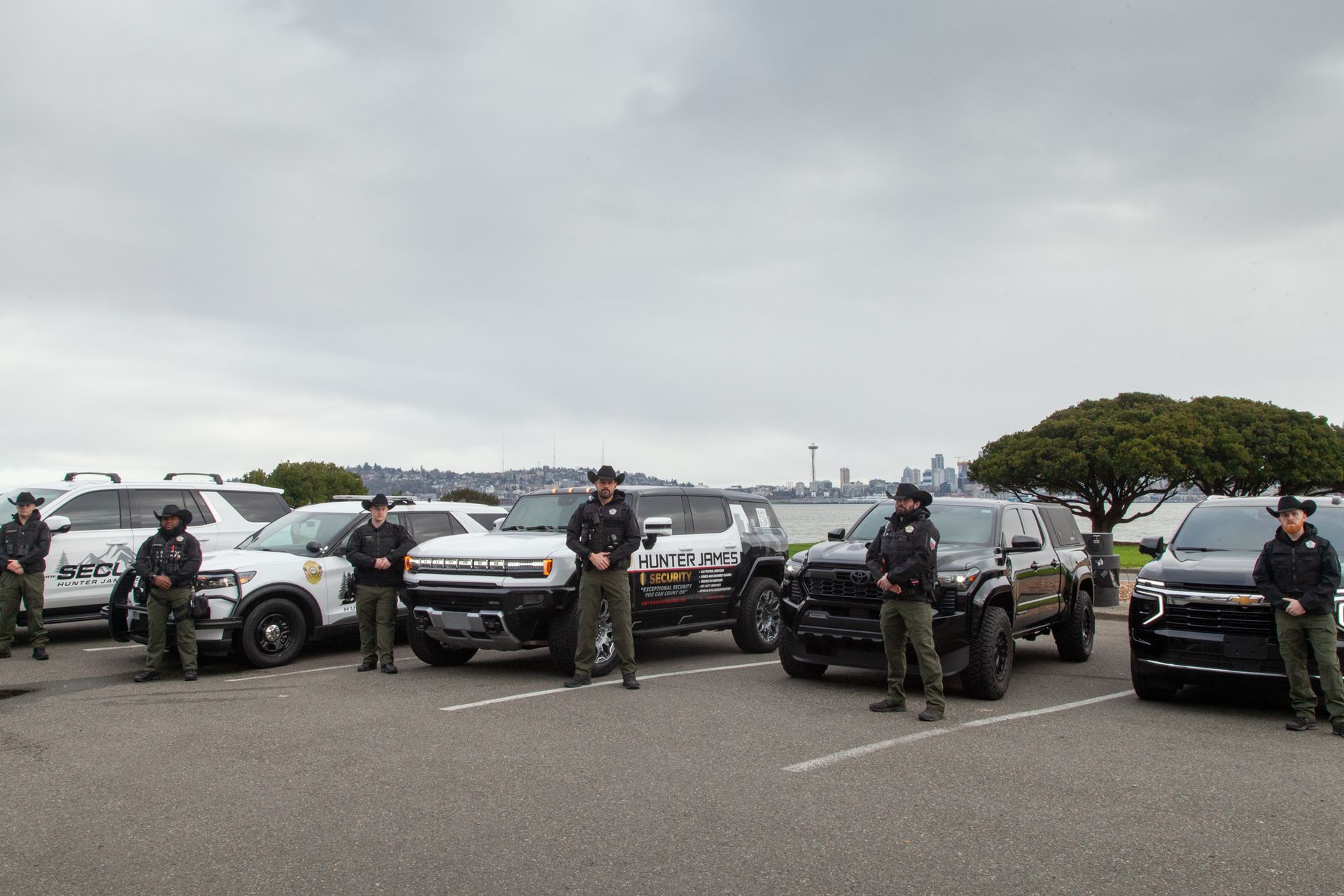 Uniformed officers stand in front of several patrol vehicles in a parking lot with a city skyline in the background.