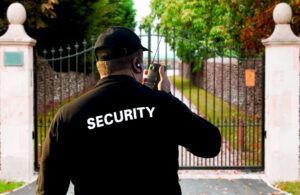 Security guard at a gate, holding a radio. The gate is black, leading to a garden.