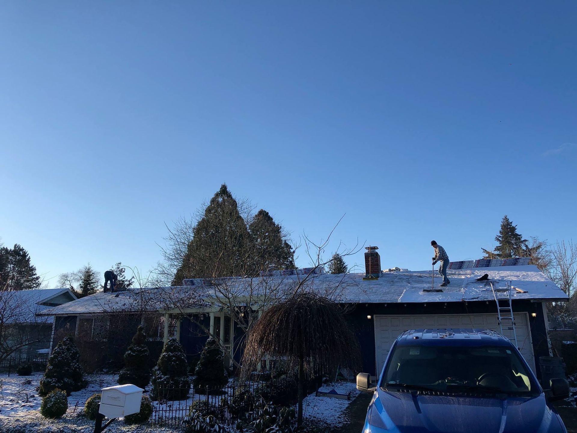 A blue car is parked in front of a house with snow on the roof.