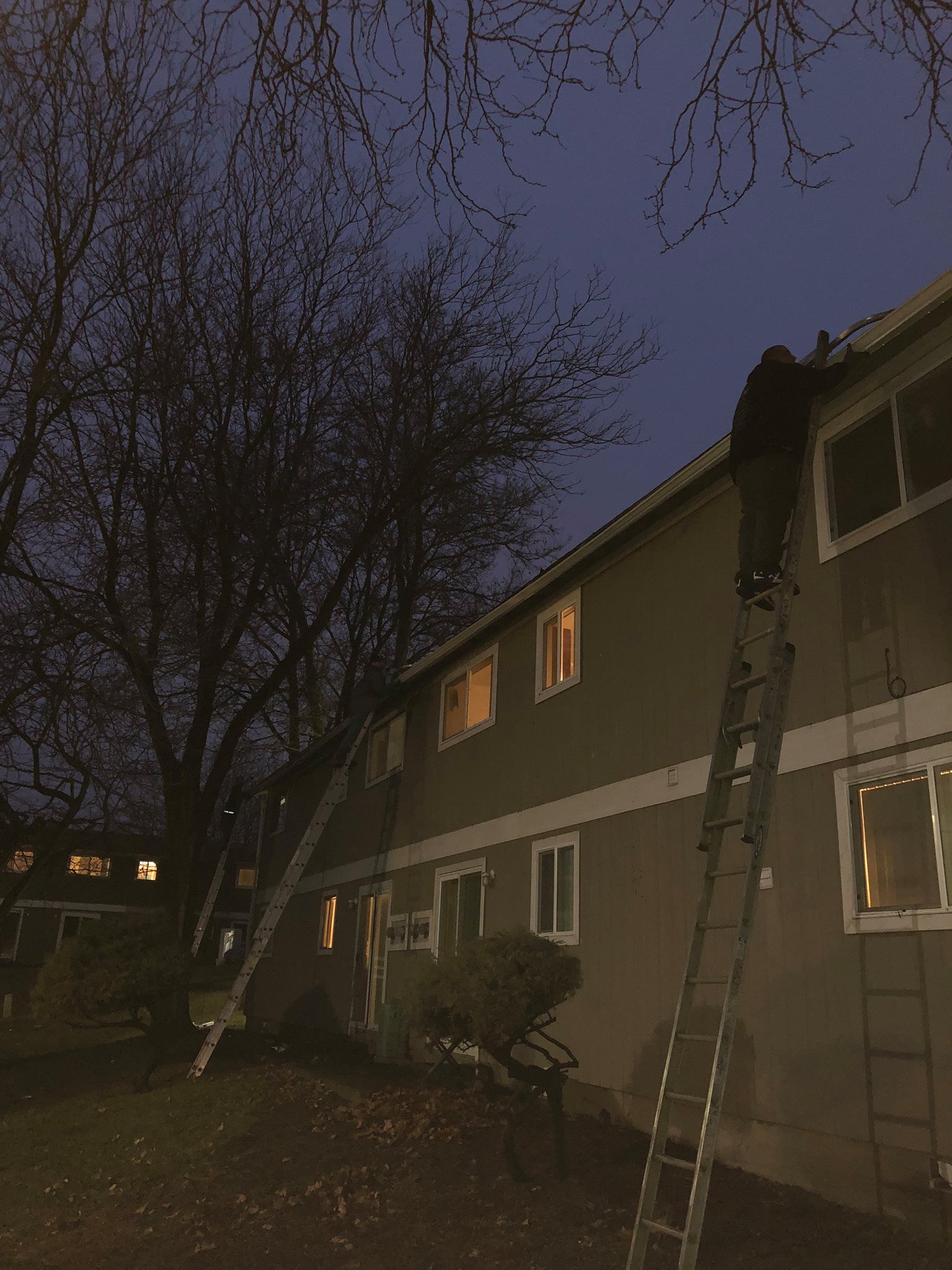 A man on a ladder is working on the roof of a house at night.