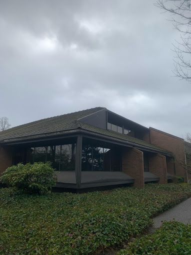A large brick building with a green roof is surrounded by bushes on a cloudy day.