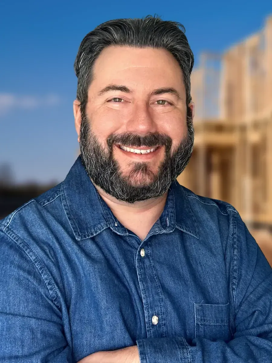 Man with a beard smiling, wearing a blue denim shirt, with construction site in the background.