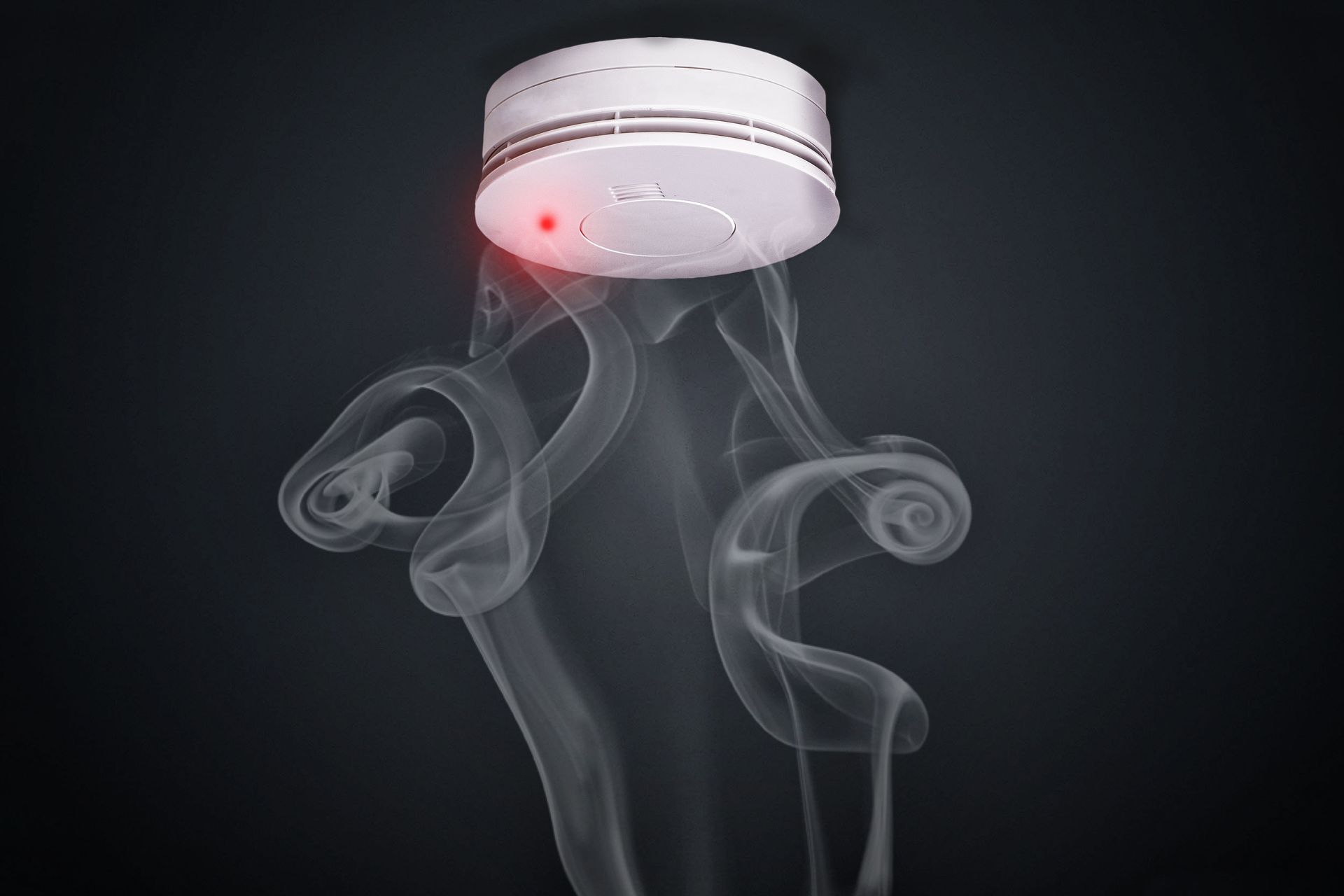 White smoke detector on dark background with a red indicator light and wisps of smoke rising beneath it