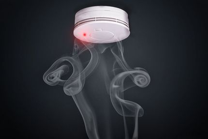 White smoke detector on dark background with a red indicator light and wisps of smoke rising beneath it
