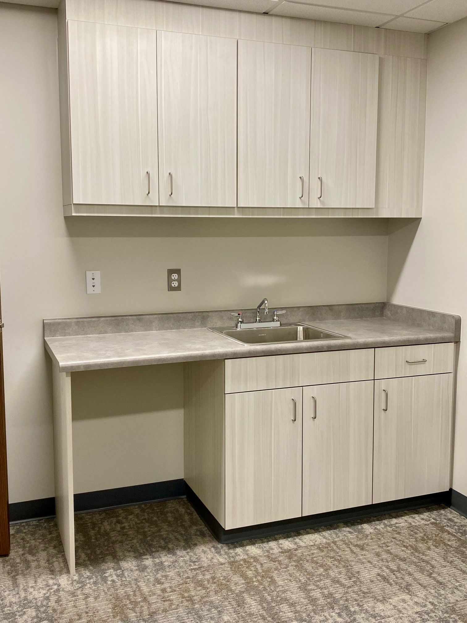 Small kitchenette with white cabinets, gray countertop, sink, and overhead storage against a beige wall.