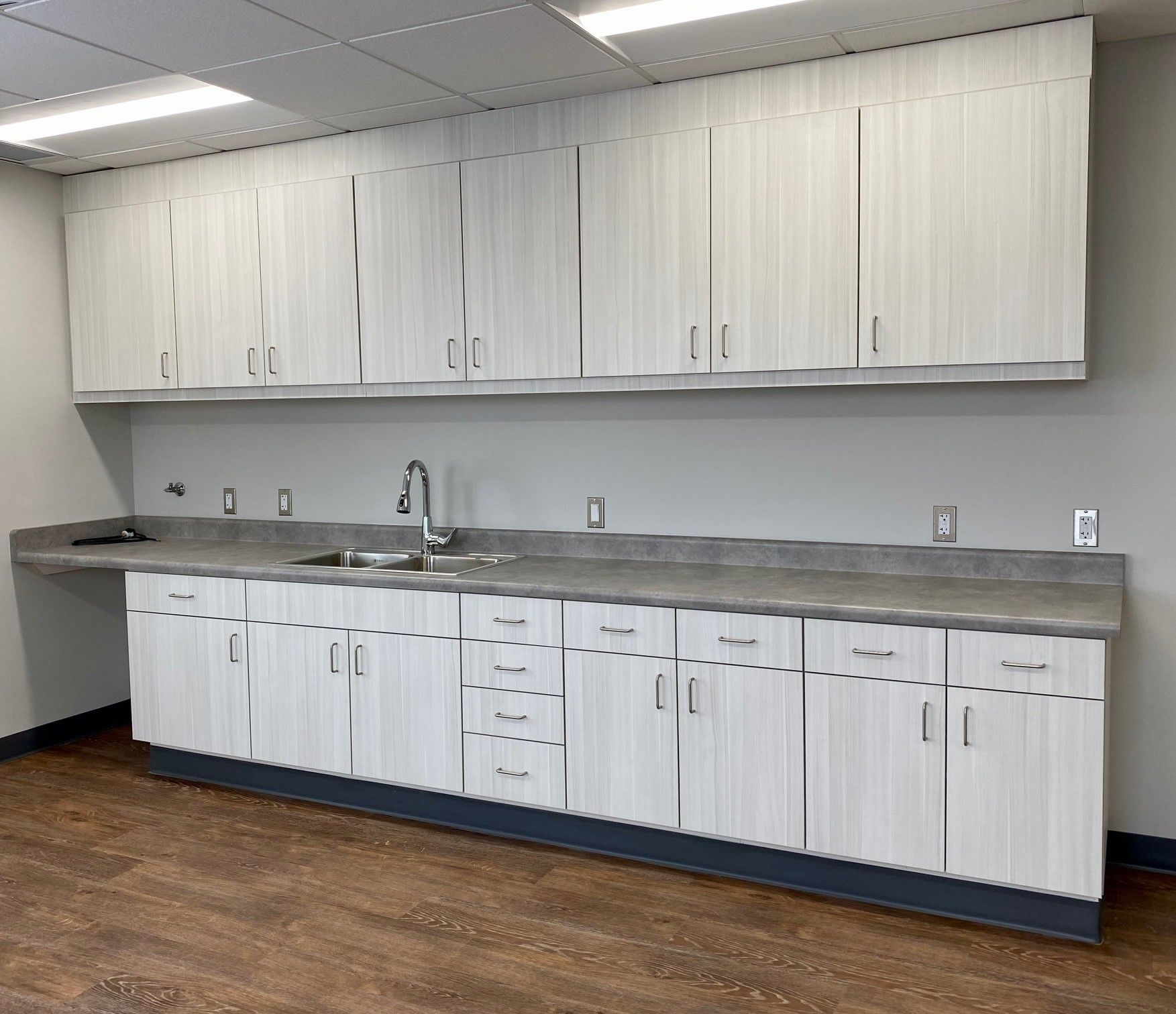 Empty kitchenette with white cabinets, gray countertop, sink, and wood floor along a wall