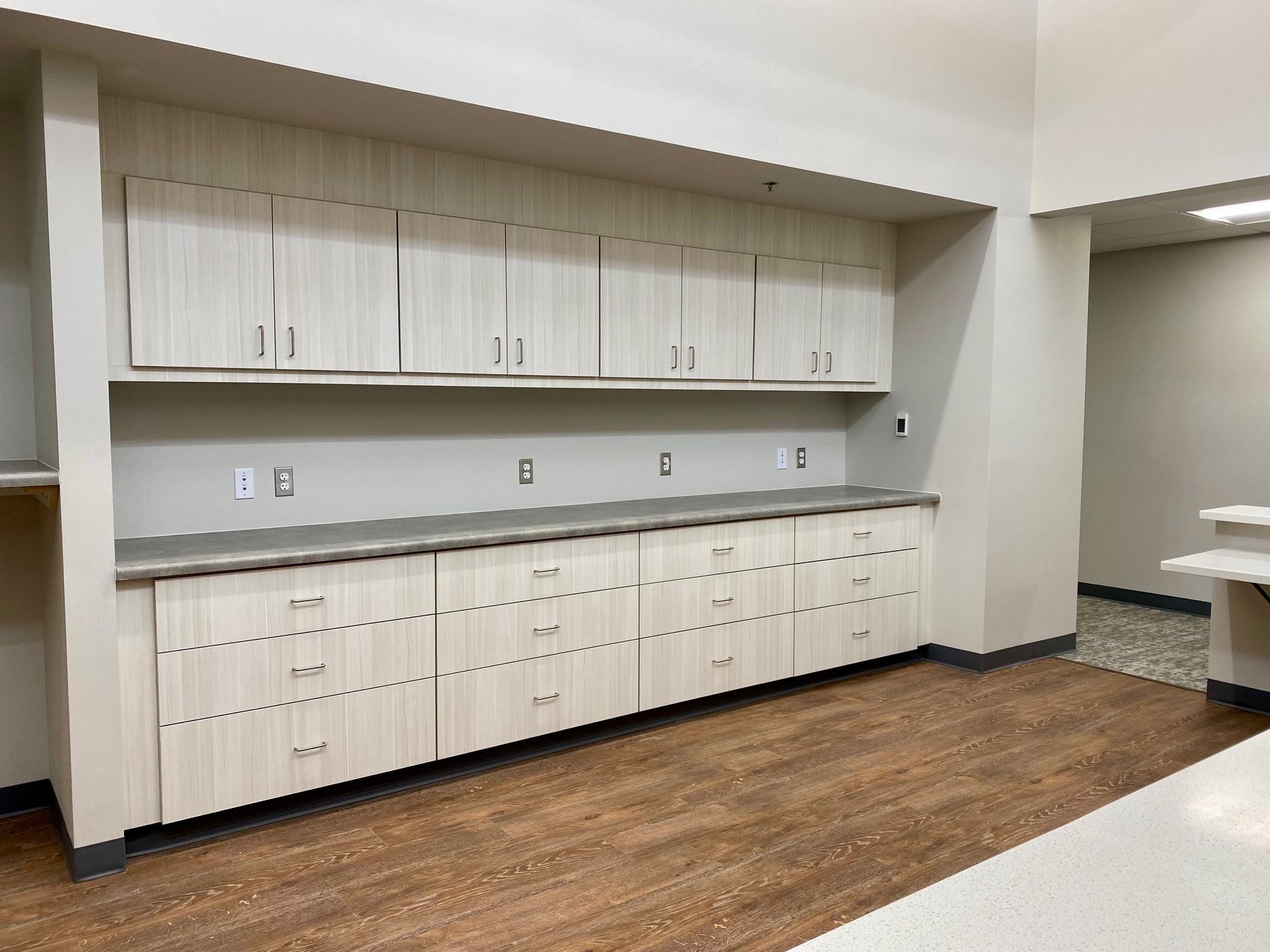Empty kitchenette with white cabinets, countertop, and wood flooring along a wall.