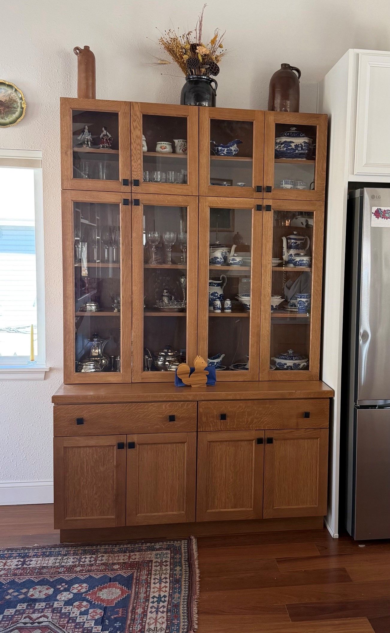 Wooden china cabinet with glass doors and shelves in a living room, with a refrigerator beside it.