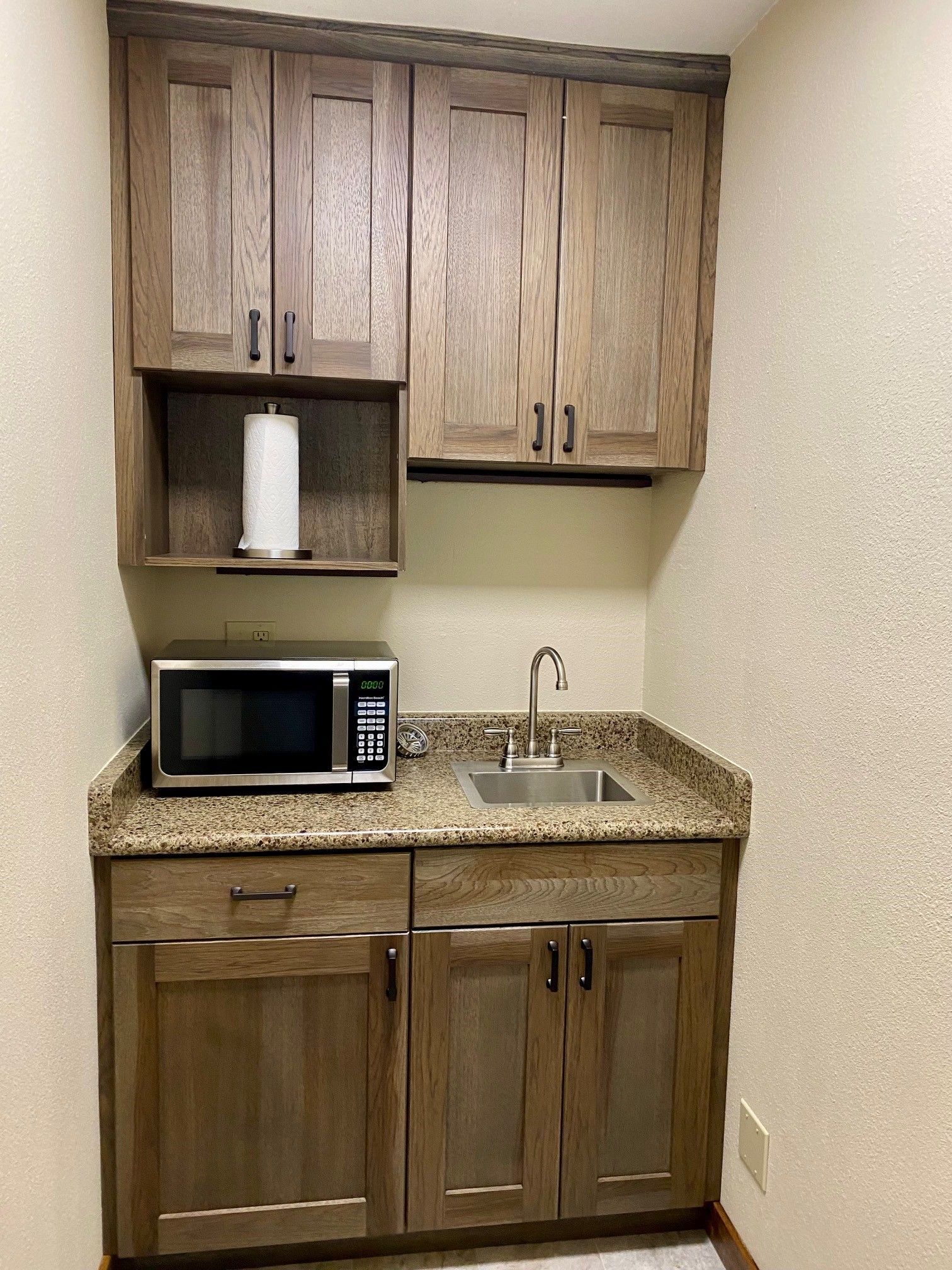 Small kitchenette with wood cabinets, granite countertop, sink, microwave, and paper towel holder.