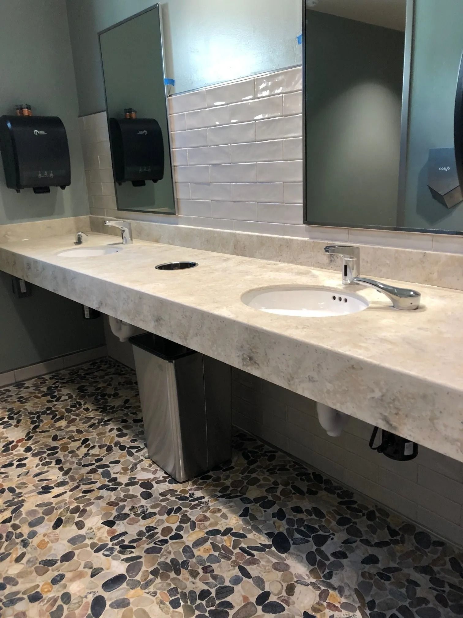 Public restroom with long stone sink counter, mirrors, soap dispensers, and speckled tile floor.