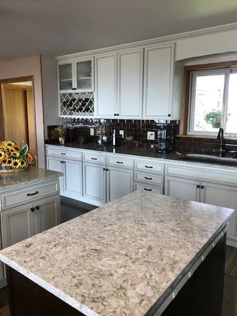 Bright kitchen with white cabinets, dark backsplash, and a large marble island by the window.