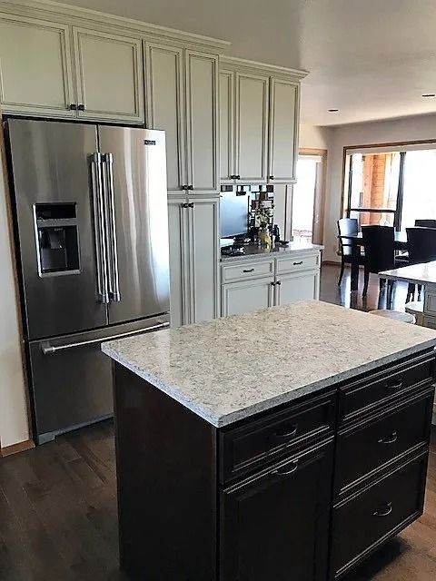 Modern kitchen with stainless steel refrigerator, light cabinets, and dark island with speckled countertop.
