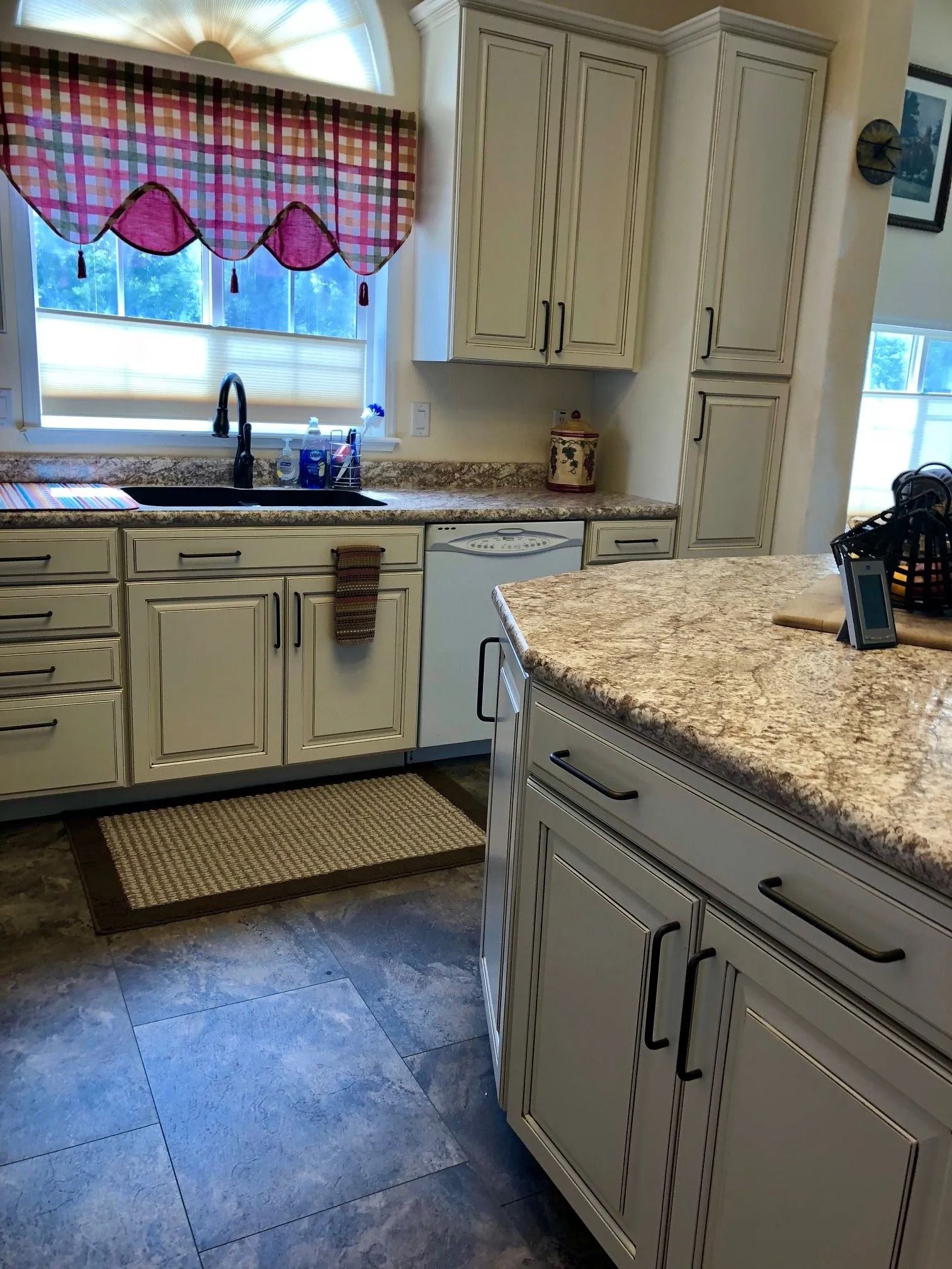 Bright kitchen with cream cabinets, granite island, tiled floor, and a sink under a window with pink scalloped curtains
