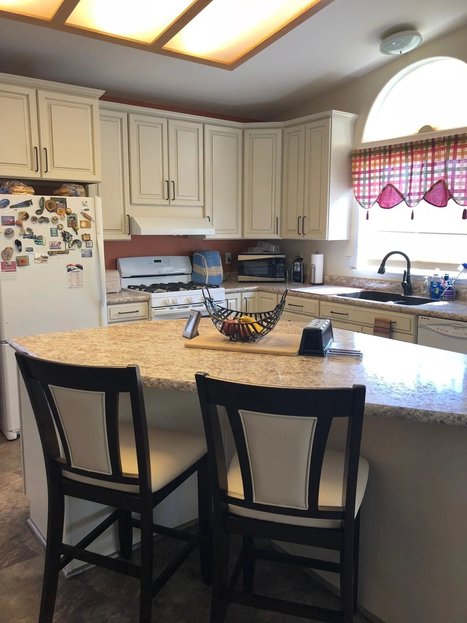 Kitchen with granite island, two black stools, cream cabinets, and a window over the sink with red curtains.