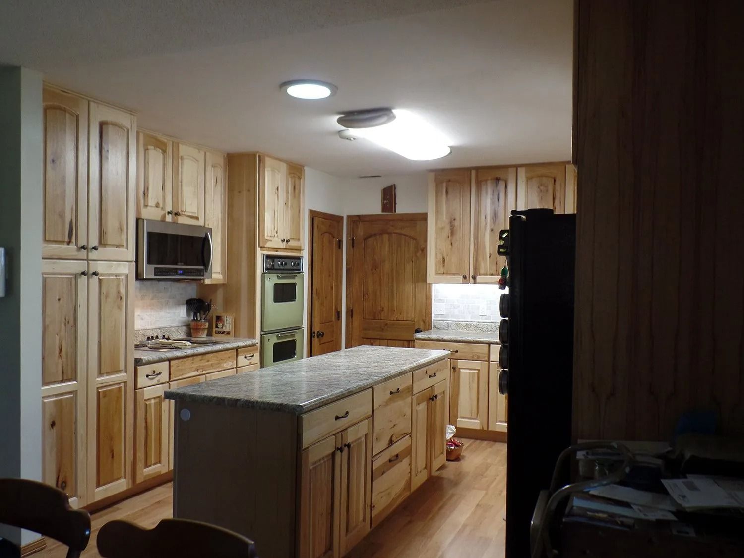 Spacious kitchen with light wood cabinets, granite island, and black refrigerator under bright ceiling lights