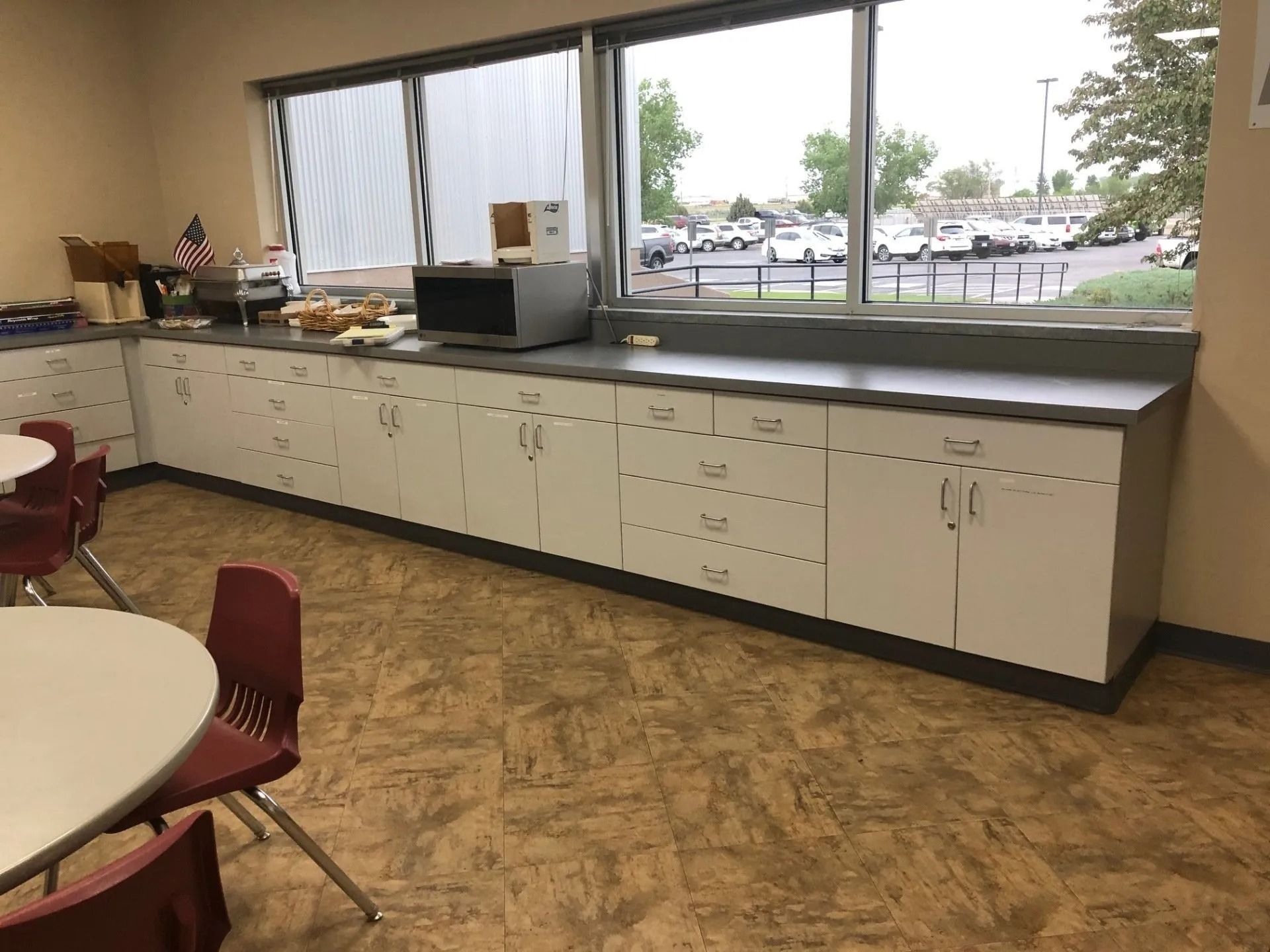 Empty cafeteria with tables, red chairs, long counter cabinets, and large windows looking out to a parking lot.