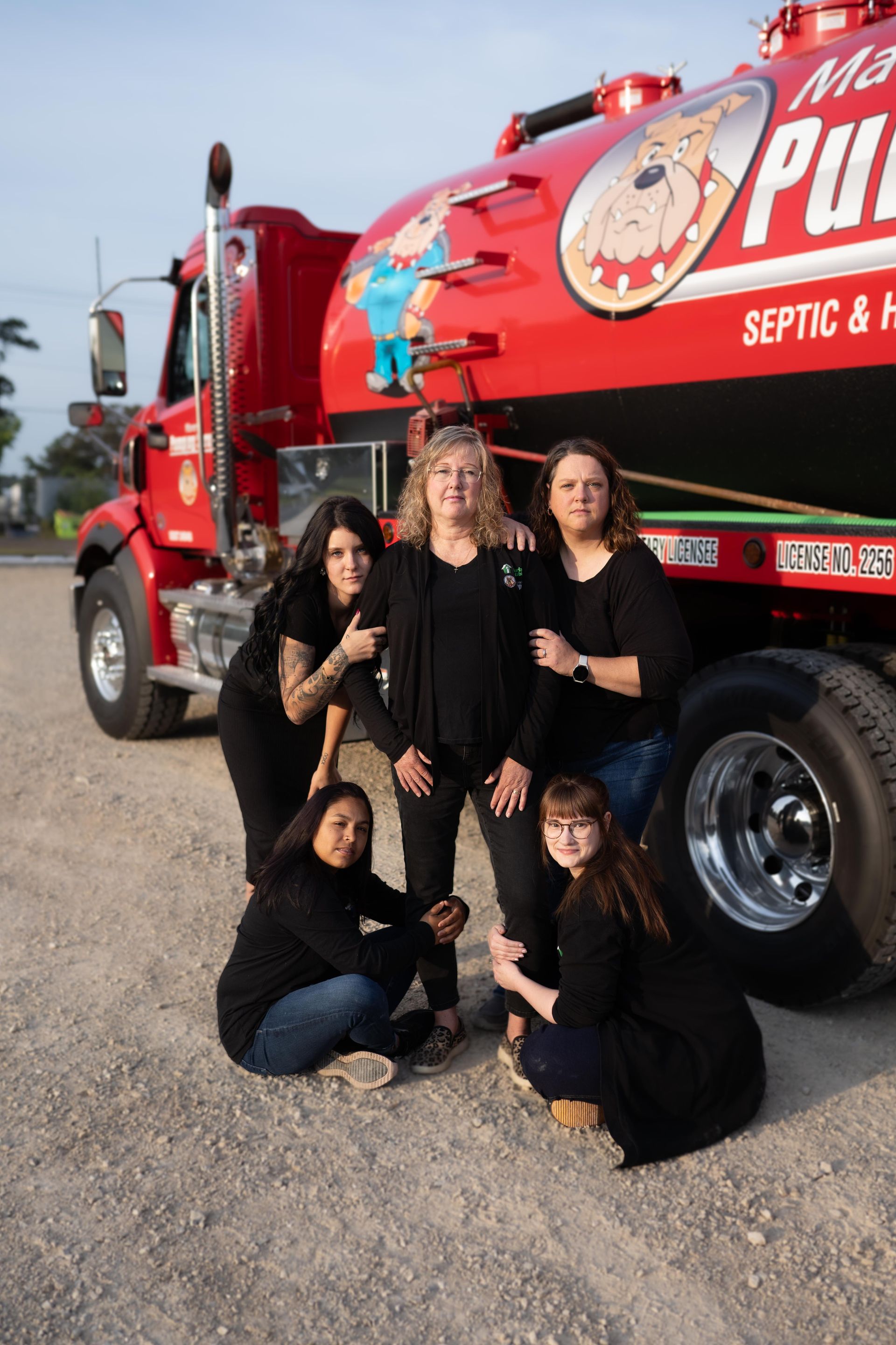 Four women pose in front of a red septic truck with a dog mascot logo.