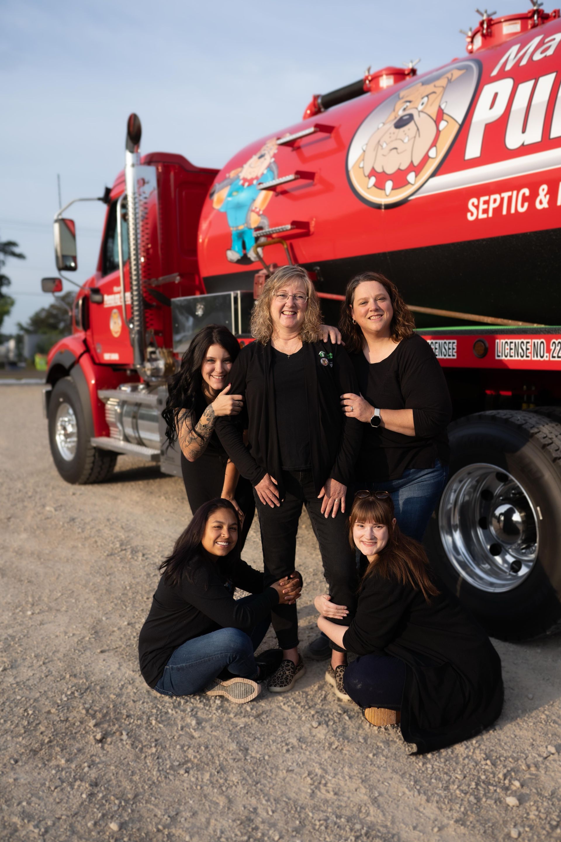 Six people pose in front of a red septic truck in a gravel lot, smiling at the camera.