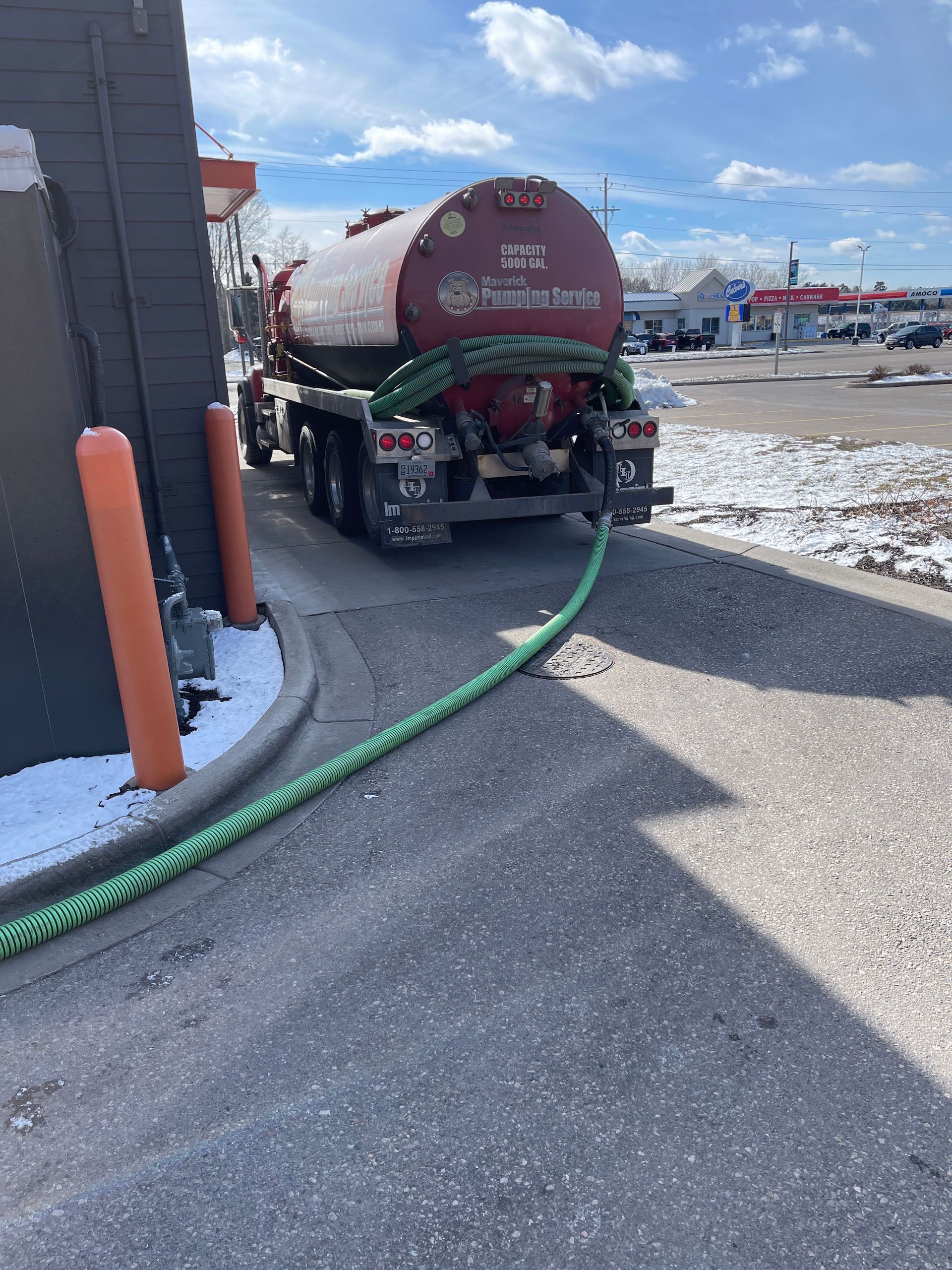 Red tanker truck parked by a building on a snowy lot, with a green hose on the ground.