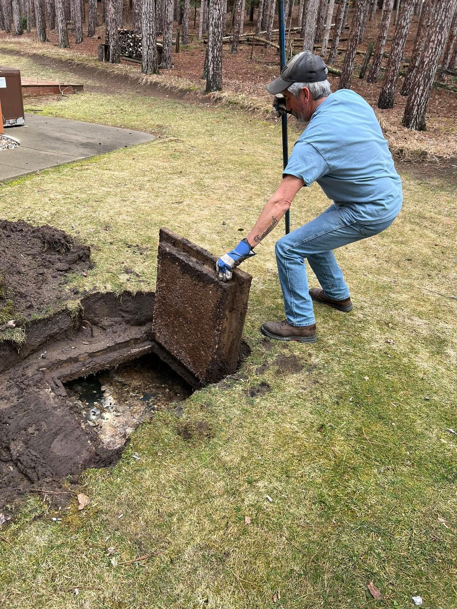 Person in blue works with a shovel at an open utility pit in a grassy yard.