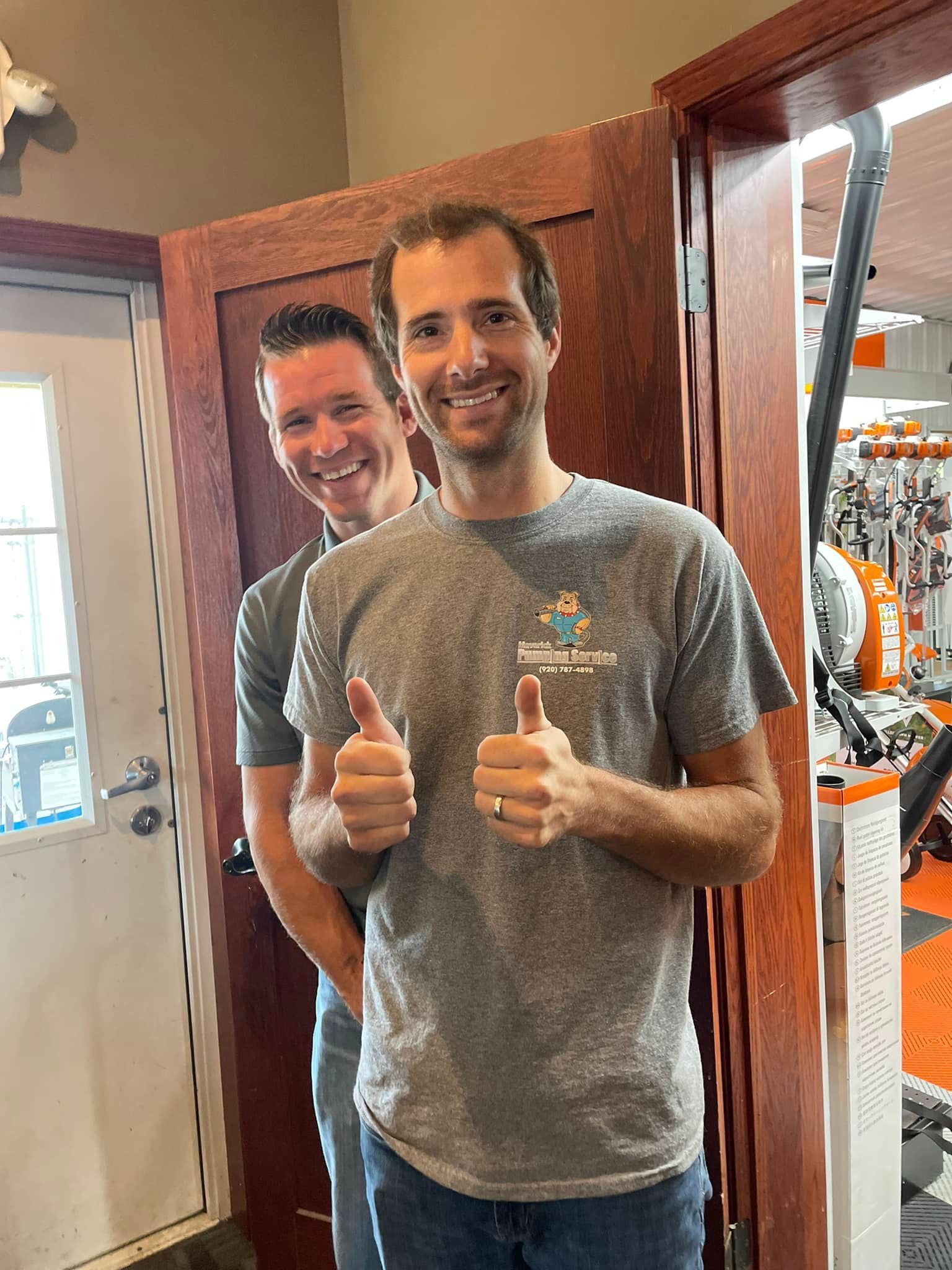 Two smiling men indoors, one in front giving two thumbs up, with orange machinery in the background.