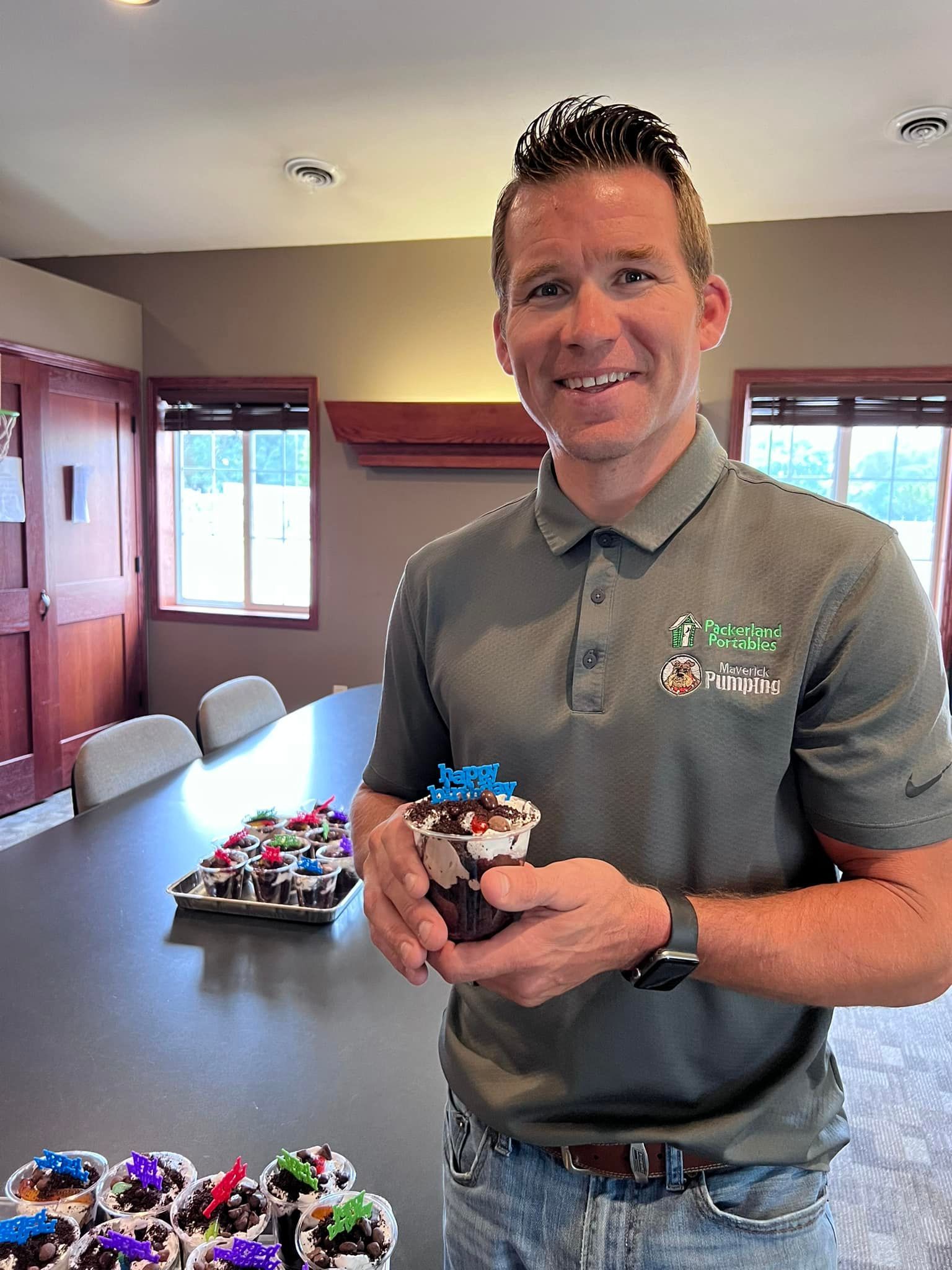 Man holding a decorated cupcake in a meeting room, with more cupcakes on a table behind him.
